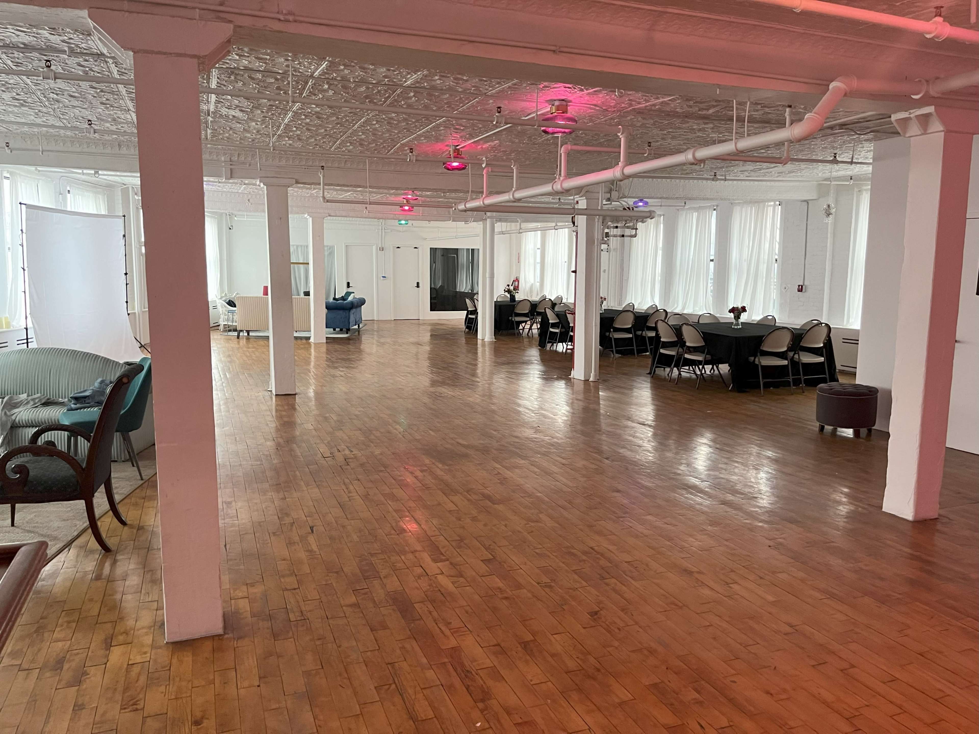 The image shows a spacious, empty room with wooden flooring, white walls, large windows, and various seating arrangements.