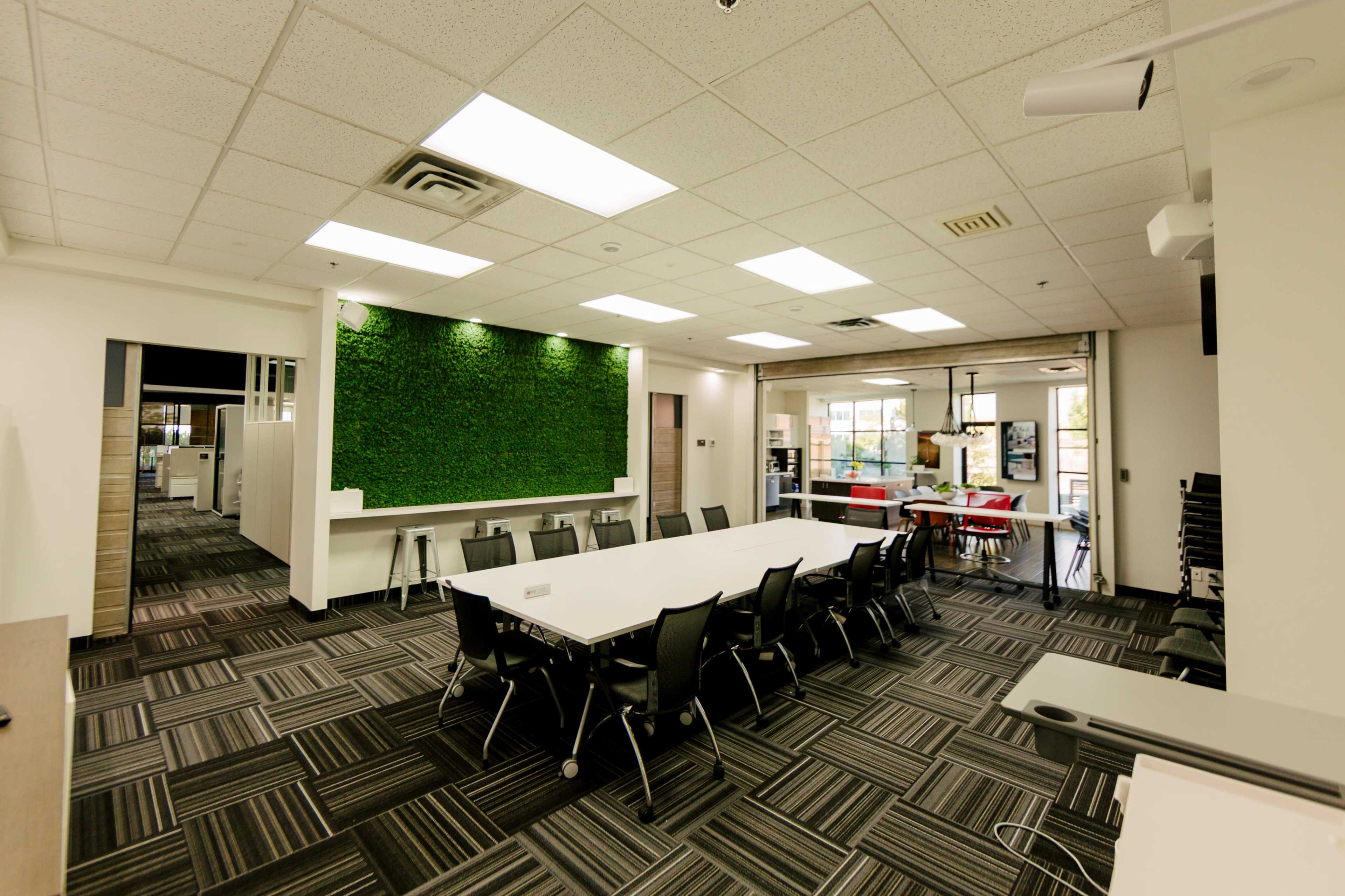 The image shows a modern conference room with a large white table, black chairs, and a green moss wall, alongside a separate casual seating area visible in the background.