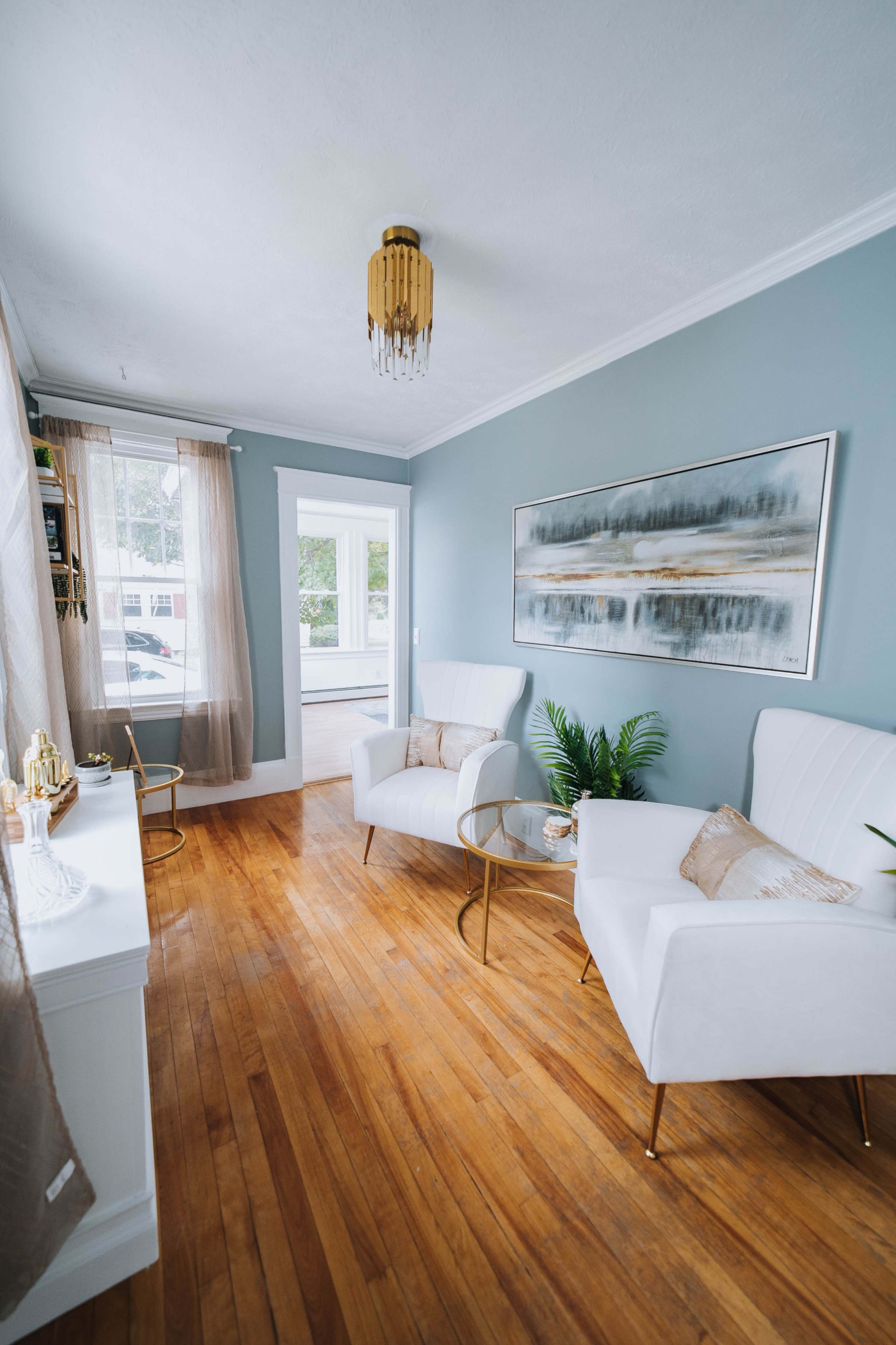 A bright living room features two white armchairs with gold legs, a small table, and light wooden floors, complemented by a large painting on the wall and natural light from a window.