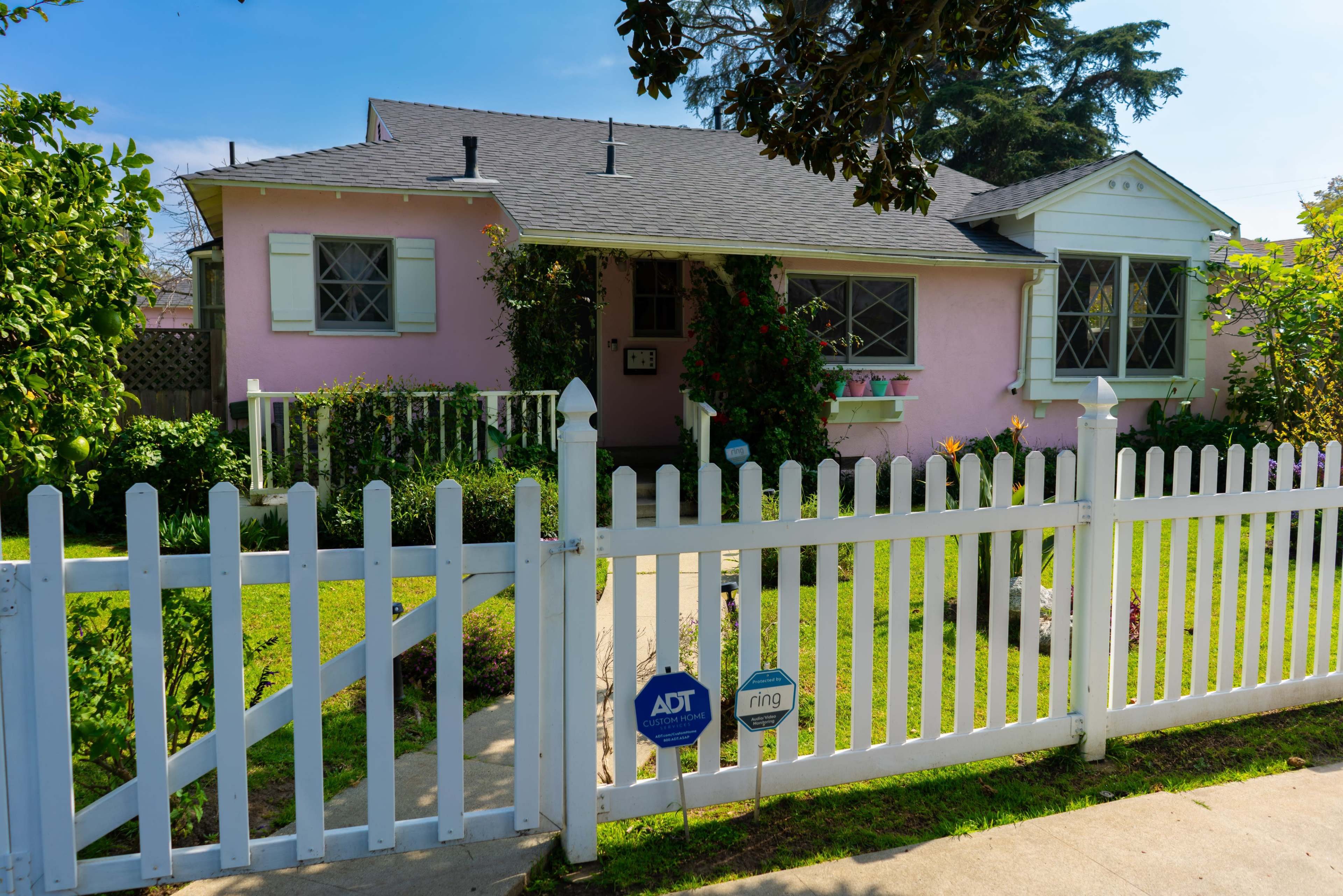 A pink house with white trim and a picket fence is surrounded by greenery and features a well-maintained front yard.
