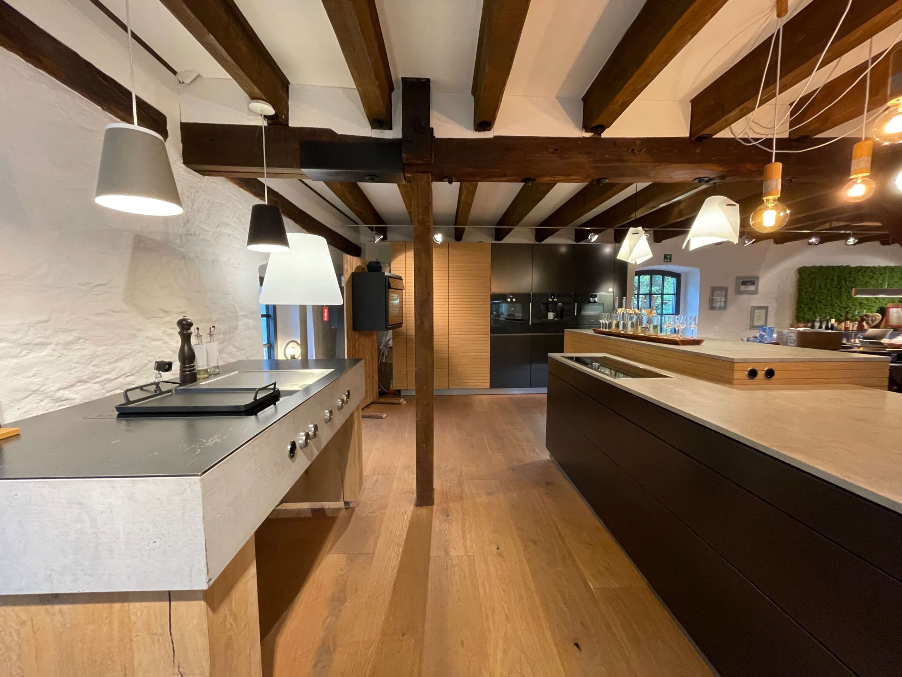 The image shows a modern kitchen with wooden beams, a concrete and black island, and various pendant lights hanging from the ceiling.