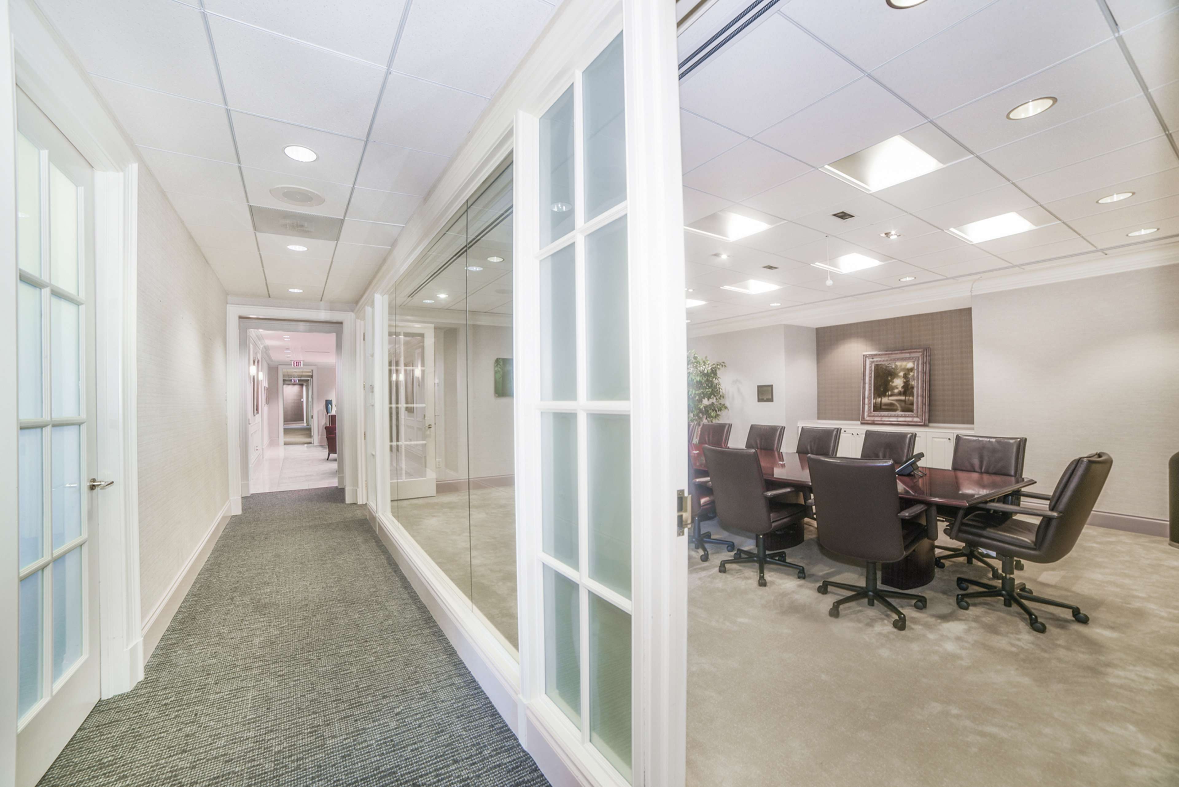 The image shows a modern office hallway leading to a glass-walled conference room with a large oval table and black chairs.