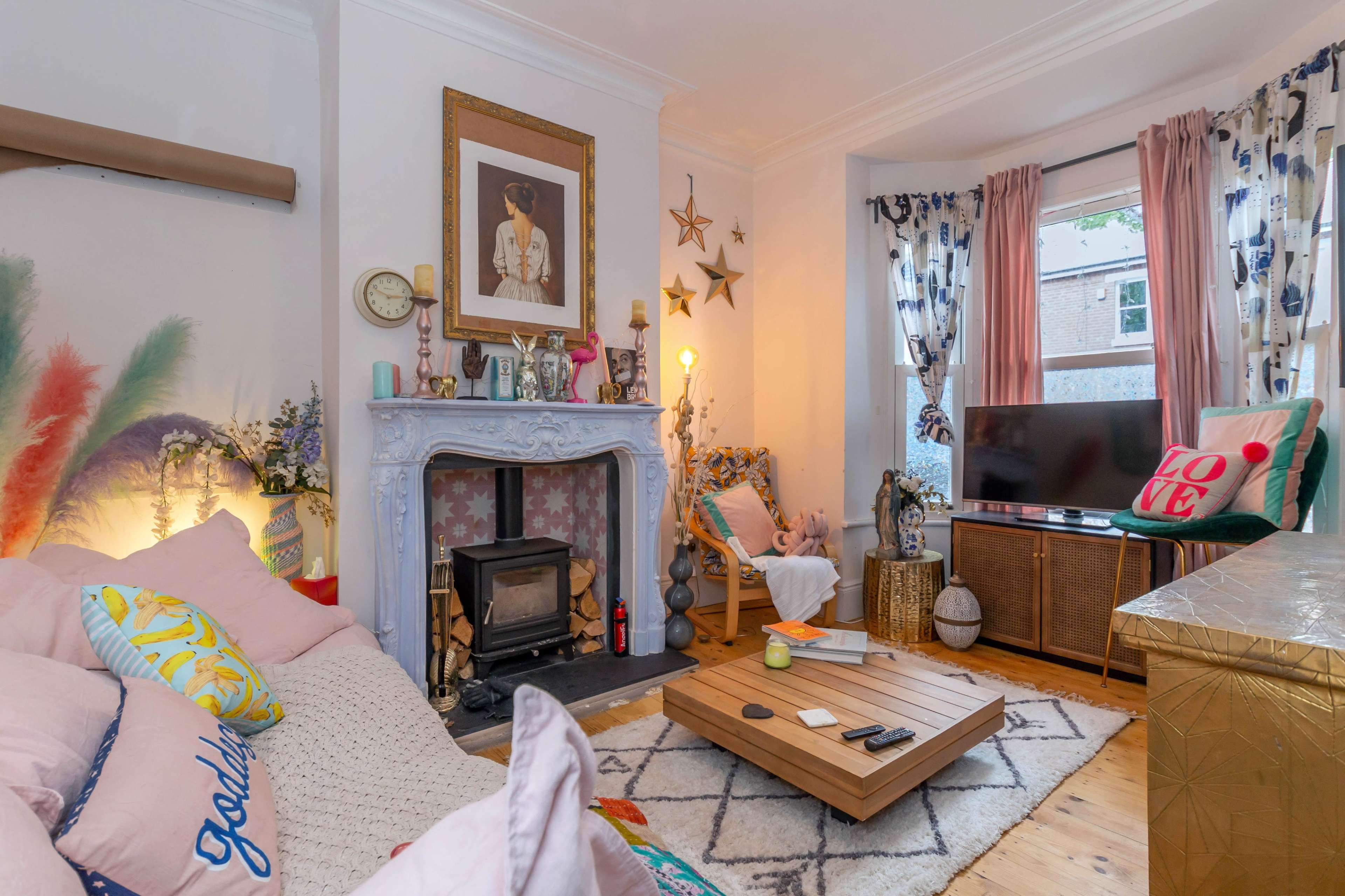 A cozy living room featuring a pastel-colored sofa with decorative pillows, a wooden coffee table, a fireplace with a blue mantel, and large windows adorned with patterned curtains.
