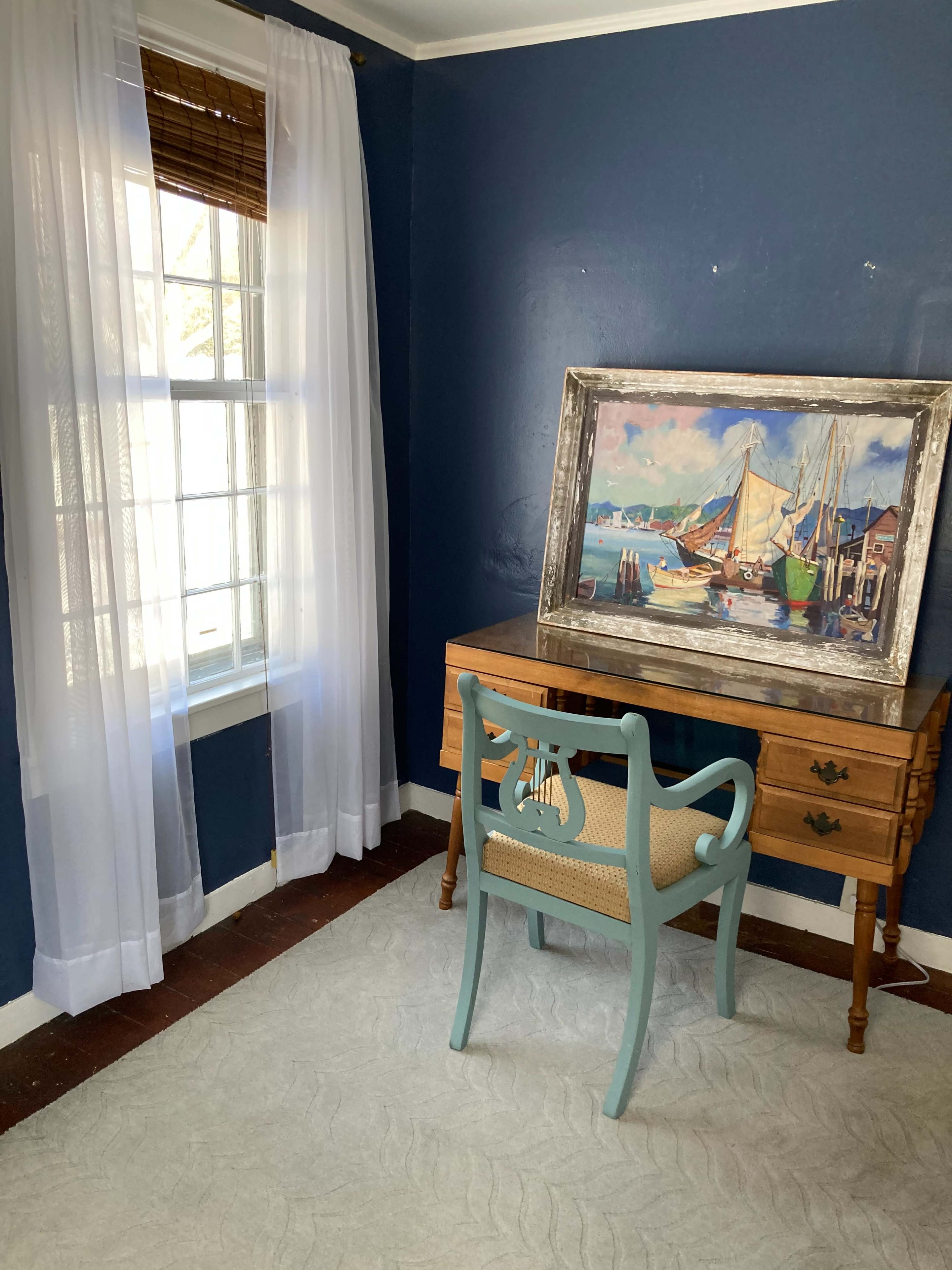 The image shows a small room with blue walls, a wooden desk, a light blue chair, and a framed painting of boats on the wall beside a window with sheer white curtains.