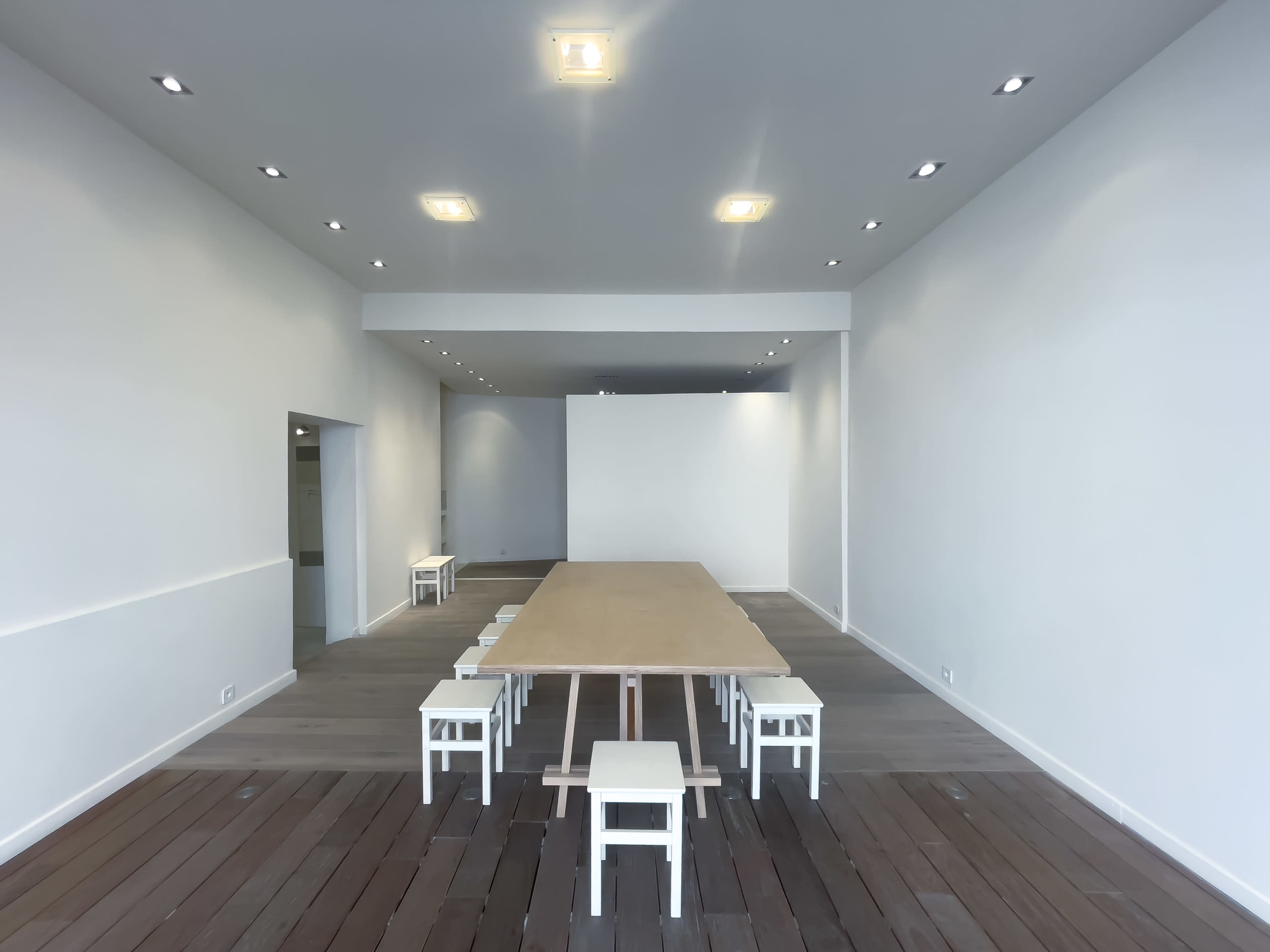 The image shows a minimalist interior of a room with a long wooden table surrounded by small white chairs, set against white walls and wooden flooring.