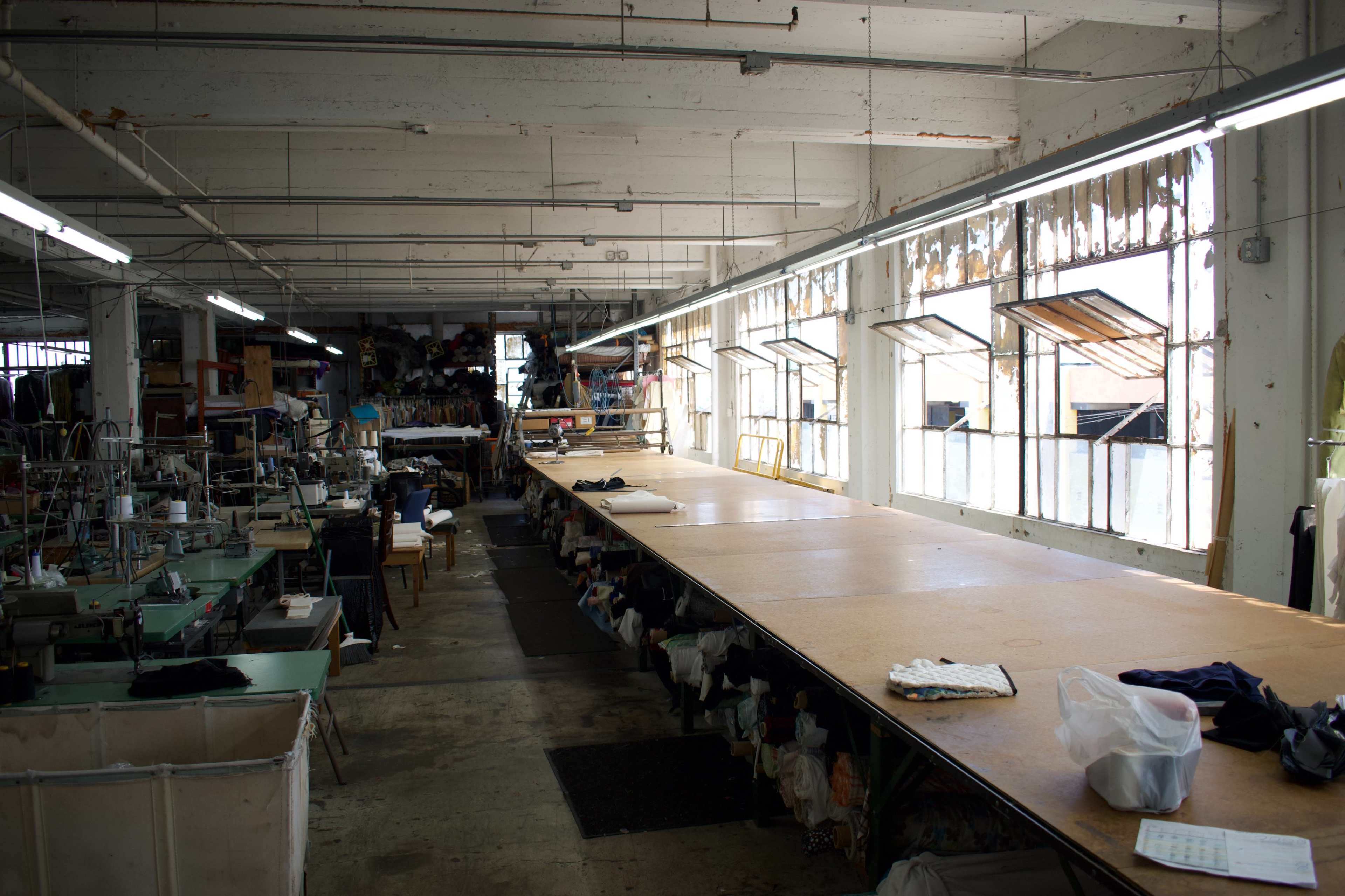 The image shows a large, open workshop space with wooden tables and sewing machines, illuminated by natural light through multiple windows.