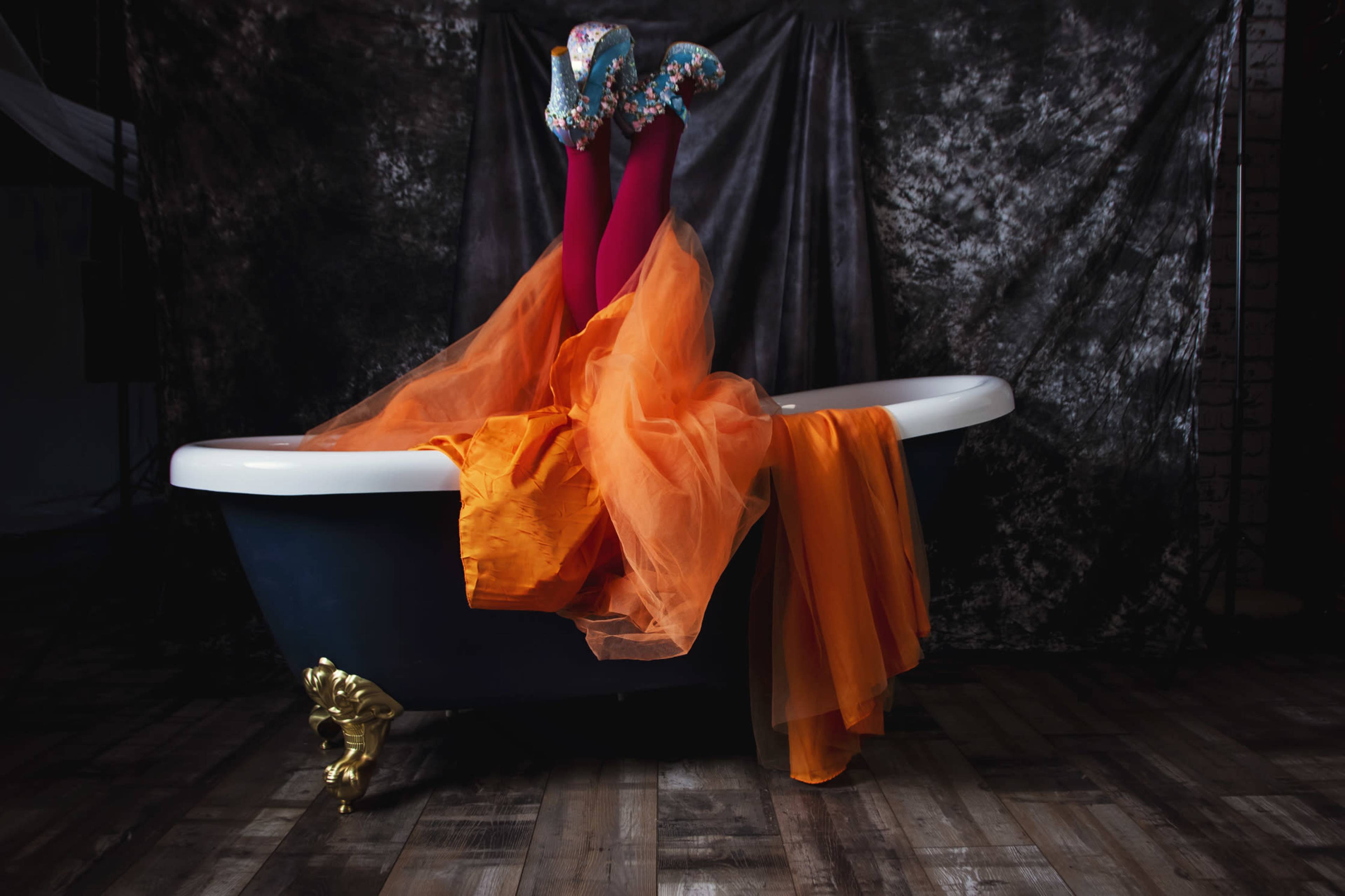 A pair of brightly clad legs with patterned shoes protrude from a vintage bathtub draped in orange fabric, set against a dark backdrop.