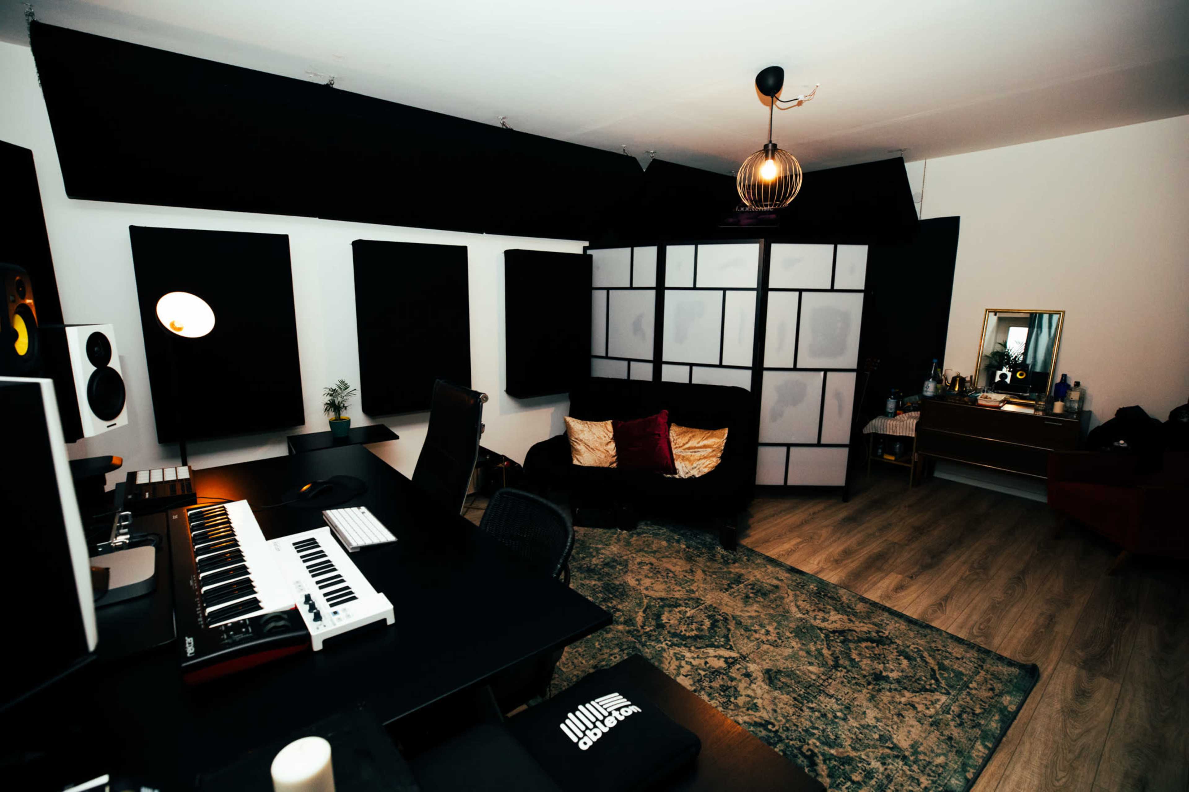 A modern music production studio featuring a keyboard on a desk, soundproof panels on the walls, and a seating area with a mirror and furniture.