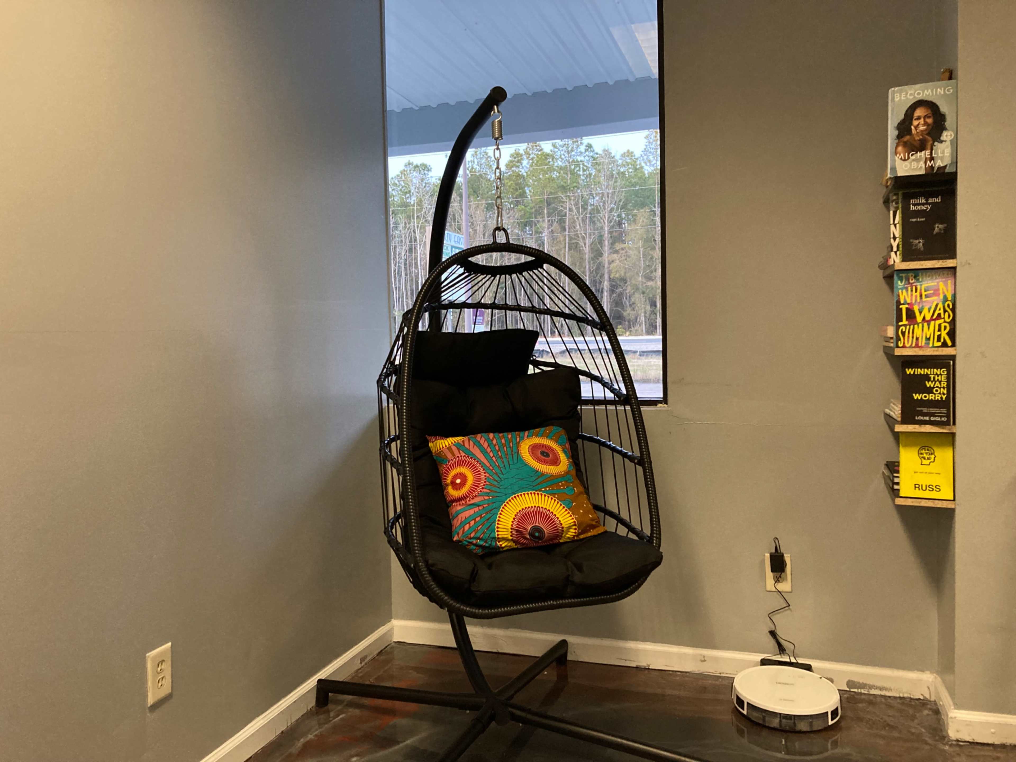 The image shows a black hanging chair with a colorful cushion, positioned near a window in a room with gray walls.