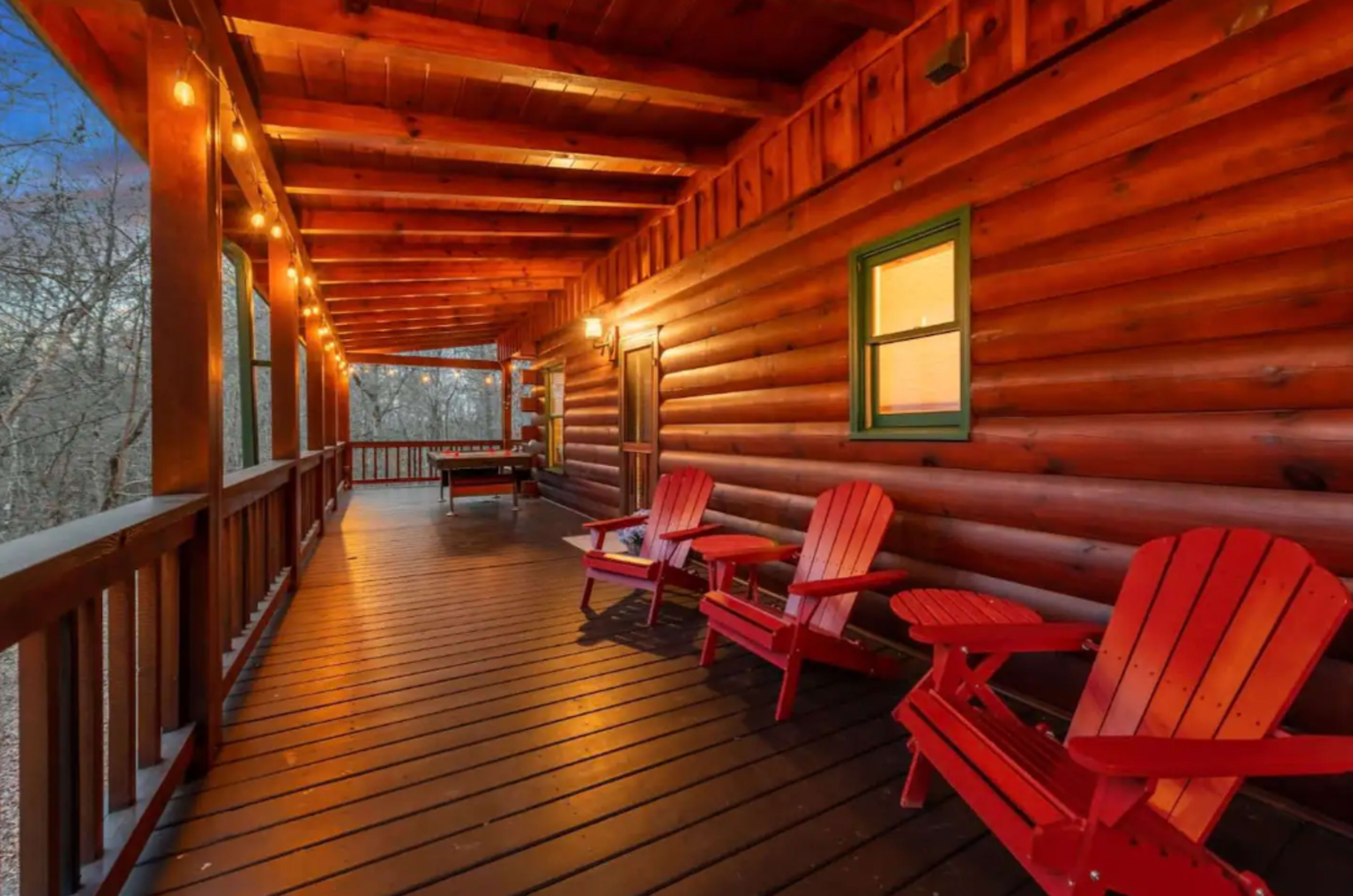 A wooden porch with red Adirondack chairs and warm string lights along the walls of a log cabin.