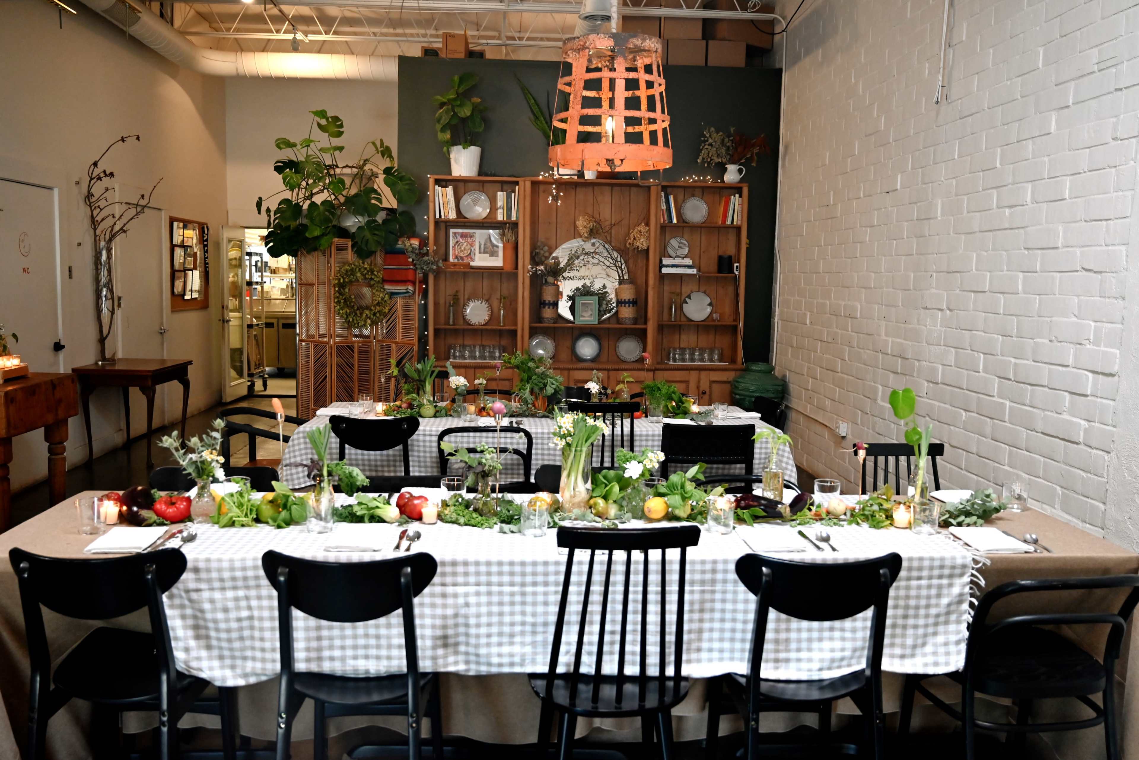 A long dining table is set with a checked tablecloth and arranged with fruits, greenery, and candles in a room featuring plants, shelves, and decorative items.