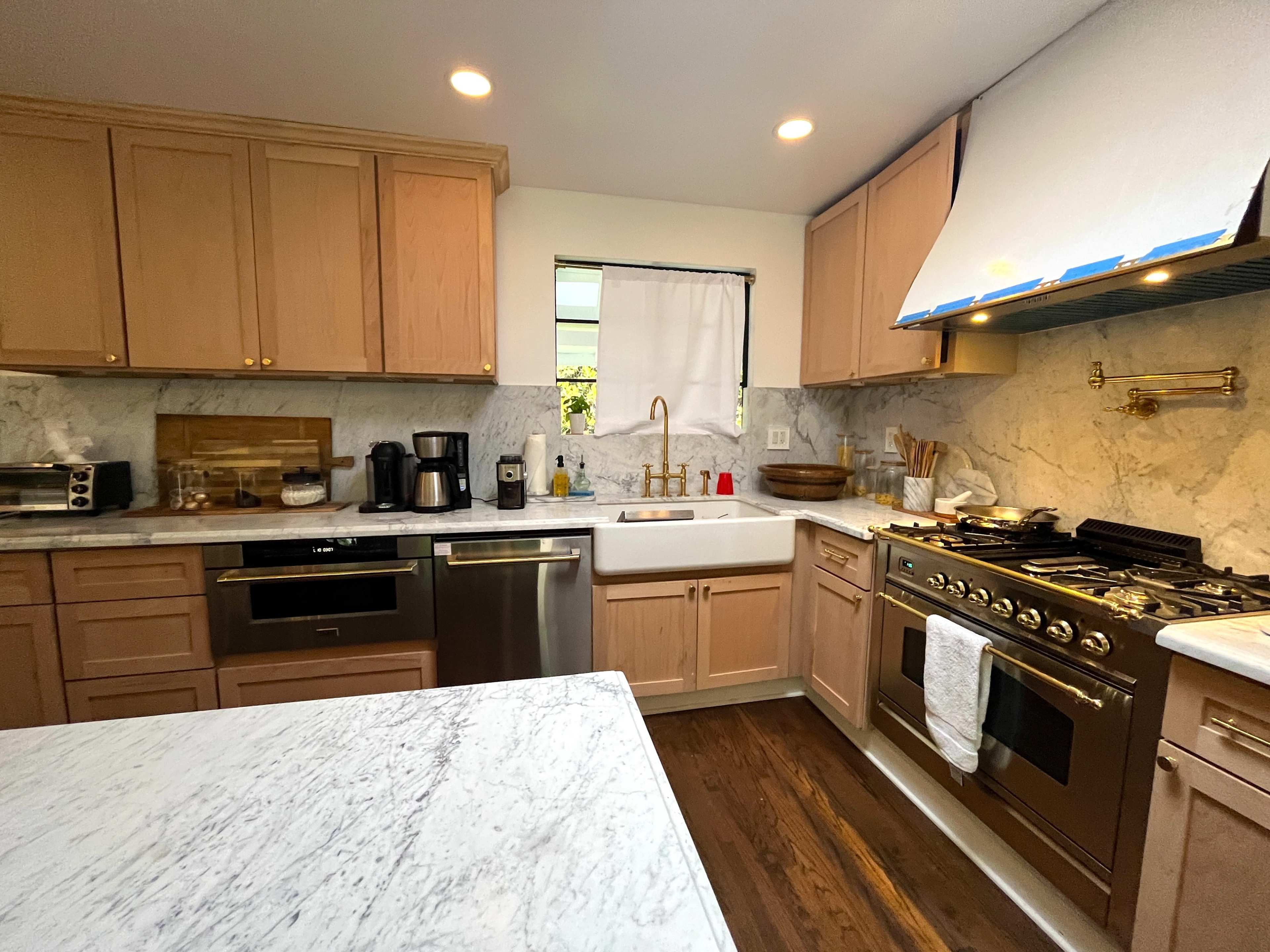 The image shows a kitchen with wooden cabinetry, a large sink, and stainless steel appliances, featuring marble countertops and a window with a natural light source.