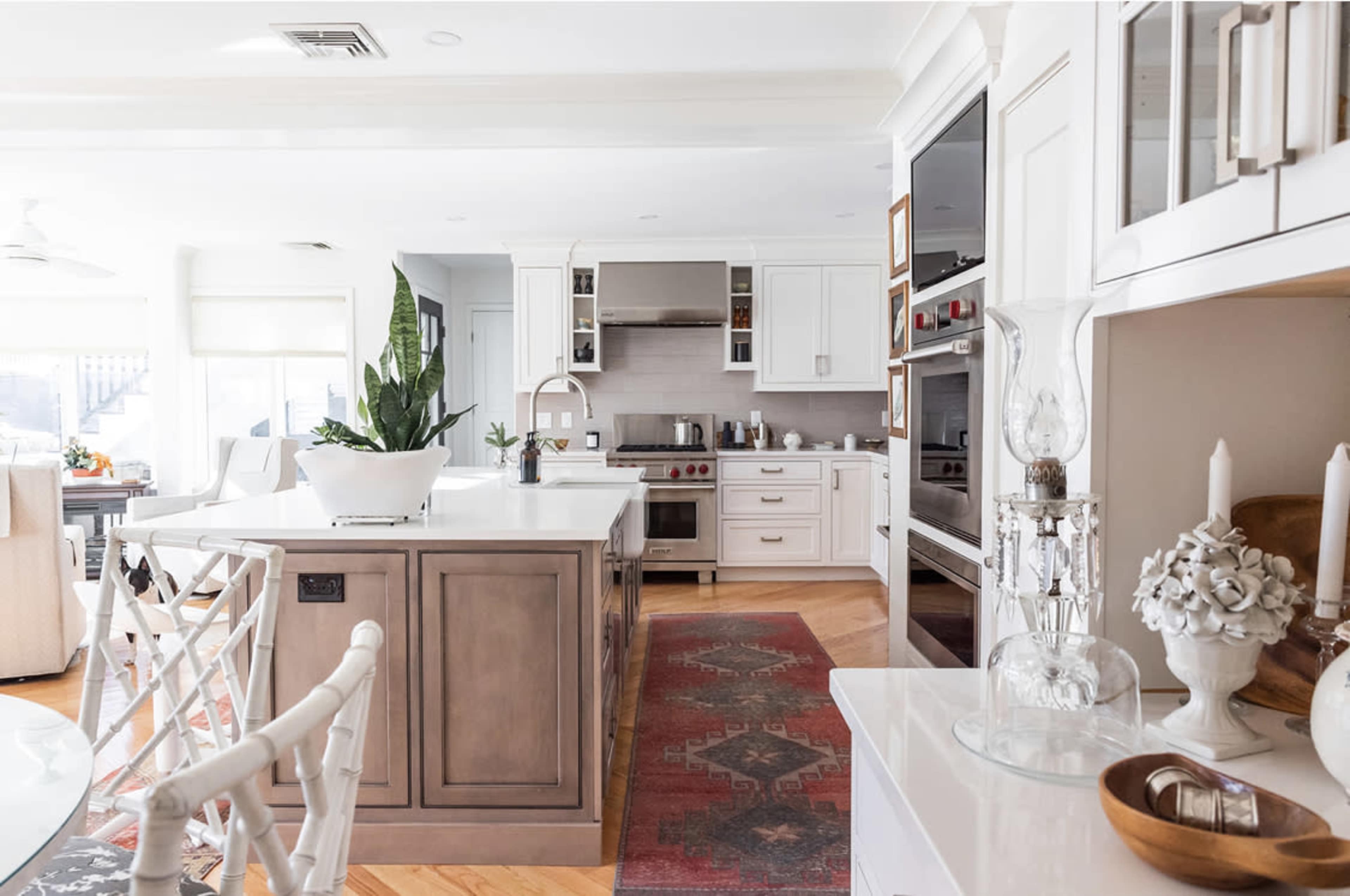 The image shows a modern kitchen with an island, featuring white cabinets, a stove, and a decorative plant, beside a dining area with a round table and upholstered chairs.