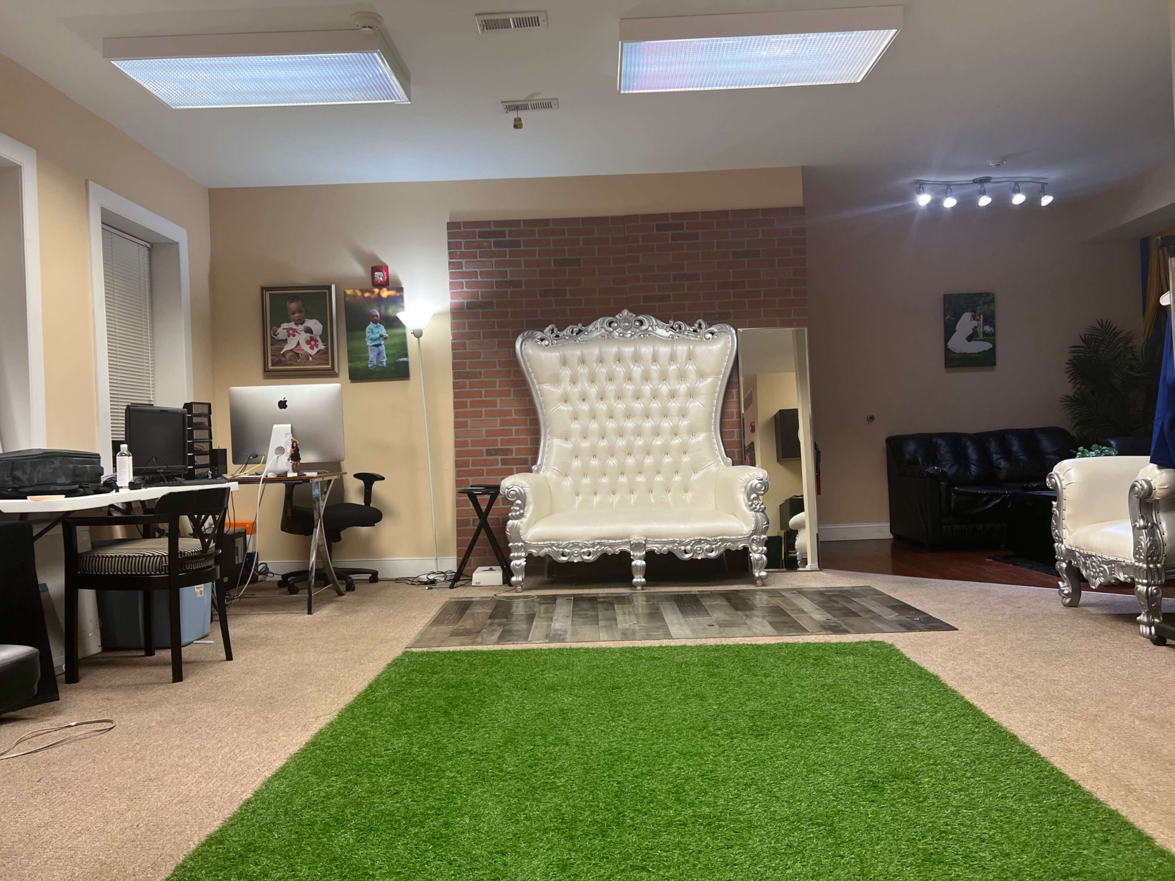 The image shows a spacious room with two ornate white chairs, a green area rug, a desk with a computer, and a cozy black couch in a well-lit environment.