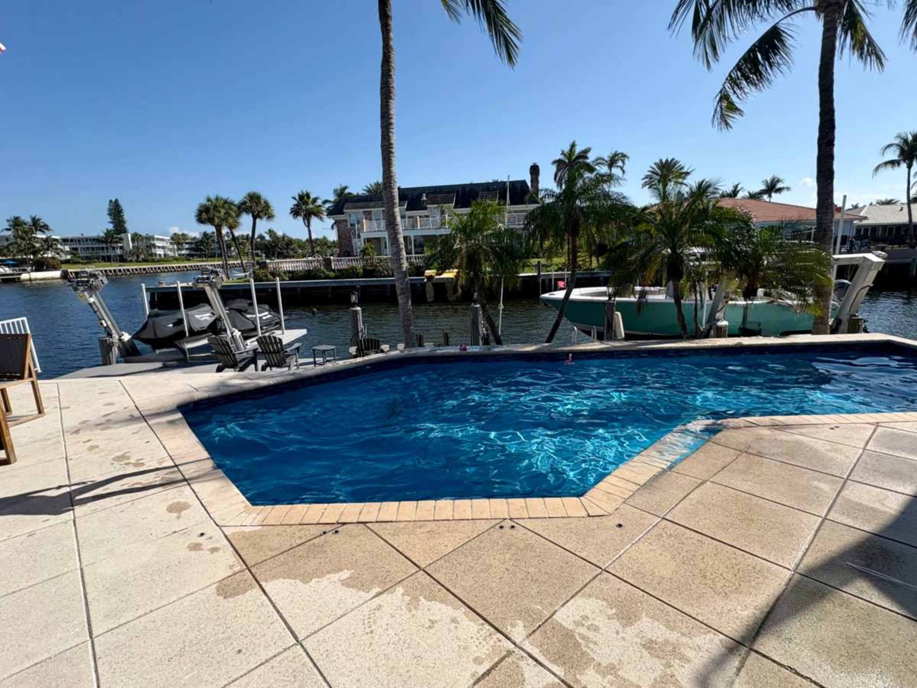 A swimming pool with clear blue water is surrounded by palm trees and overlooks a canal with parked boats.