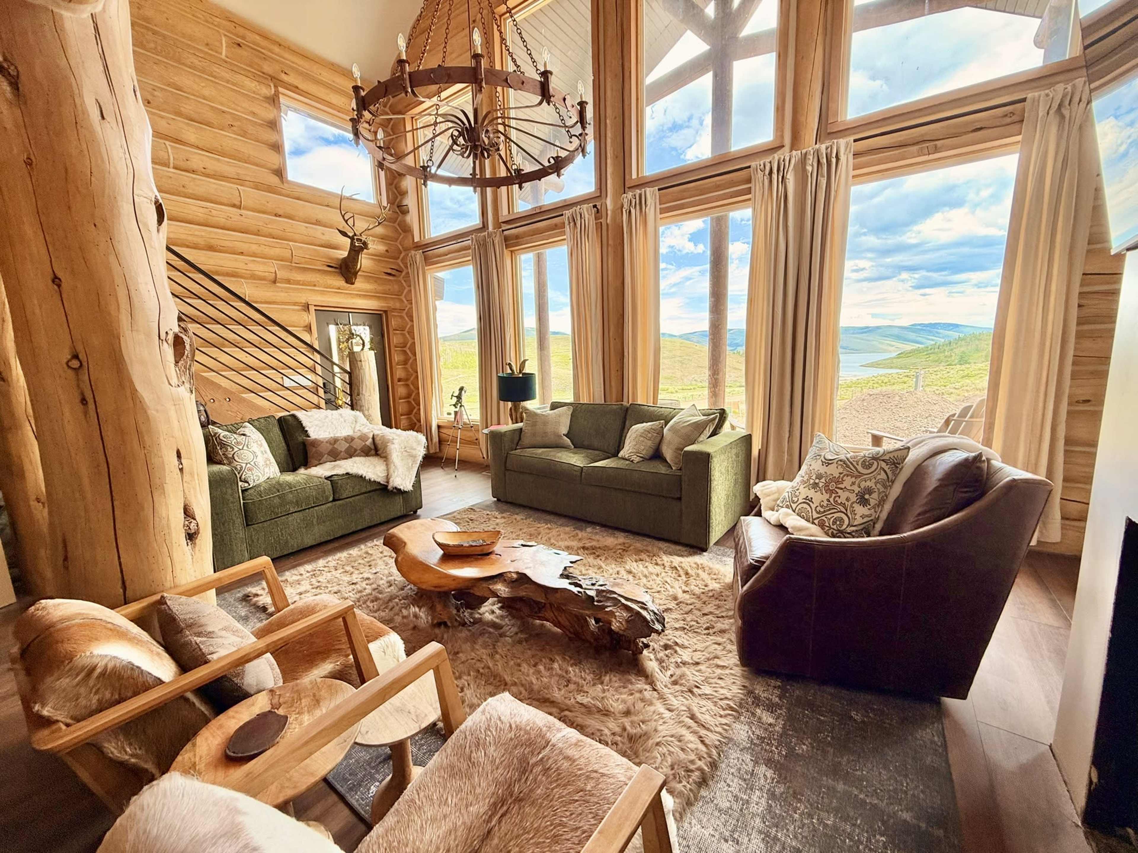 The image features a spacious, rustic living room with large windows showcasing a scenic view and furnished with multiple seating areas.