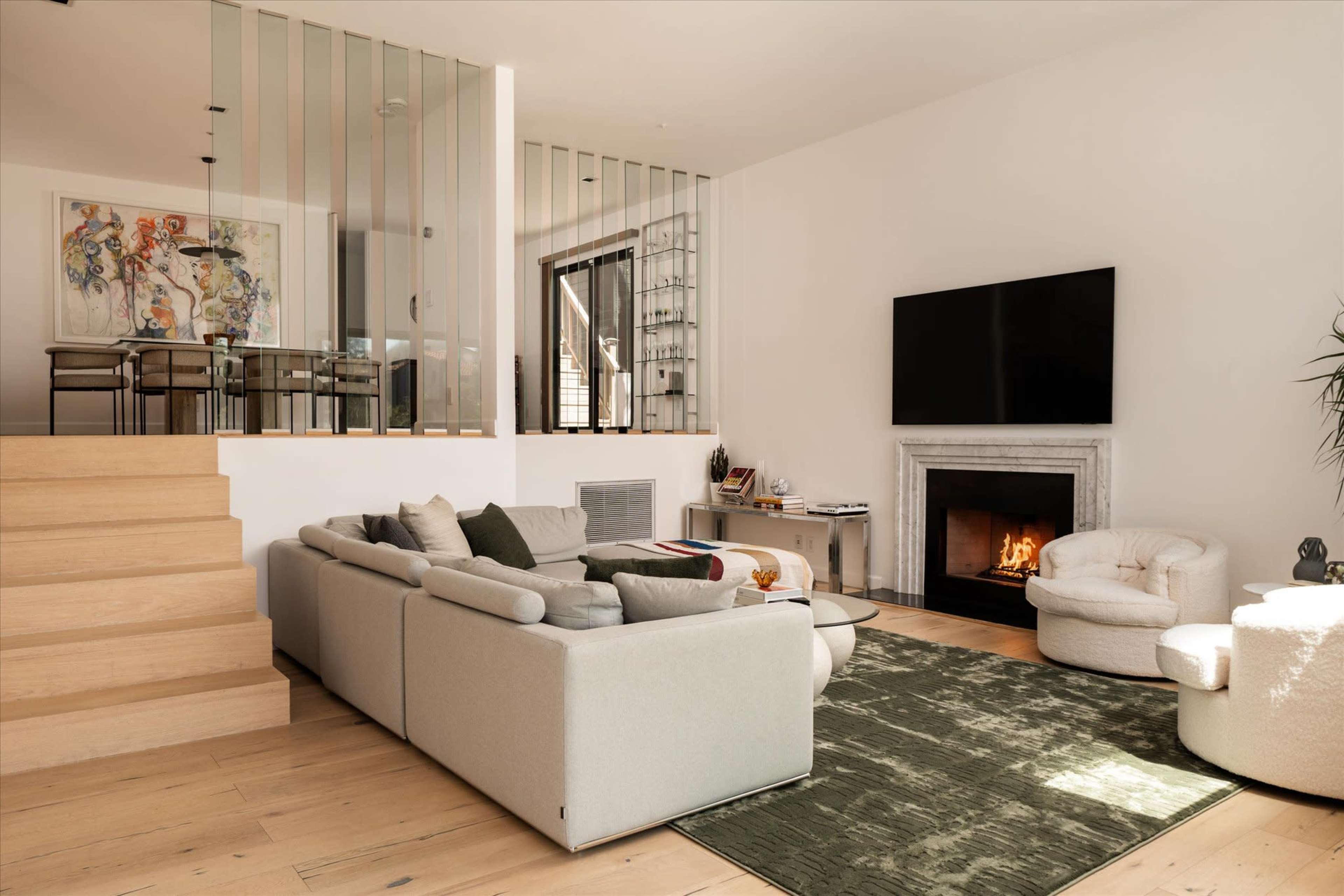 A modern living room features a large gray sectional couch, a fireplace, and a staircase leading to an open dining area with glass partitions.