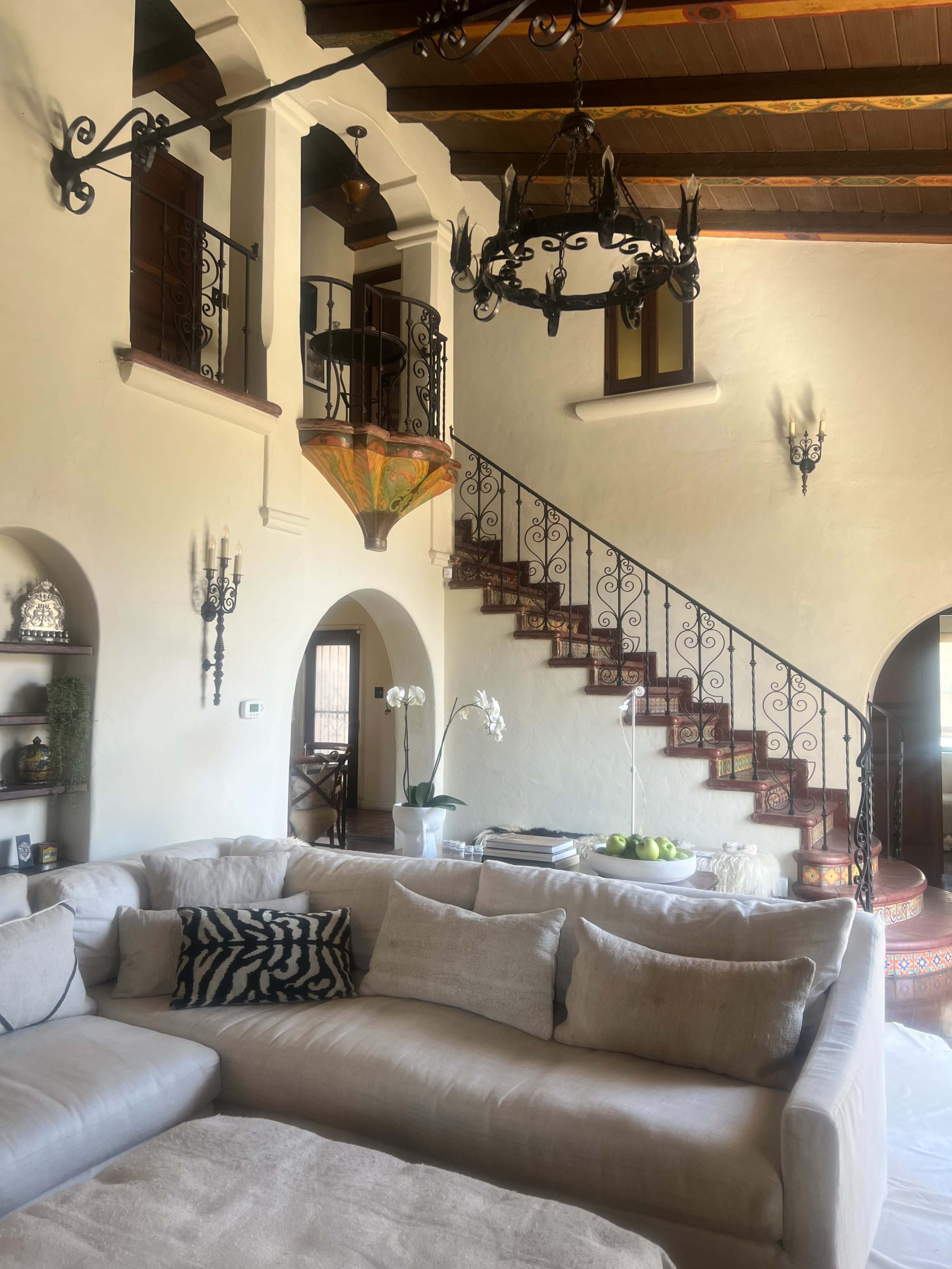 The image shows a spacious living area with a large light-colored sofa, an ornate staircase with wrought iron railings, and a decorative chandelier.