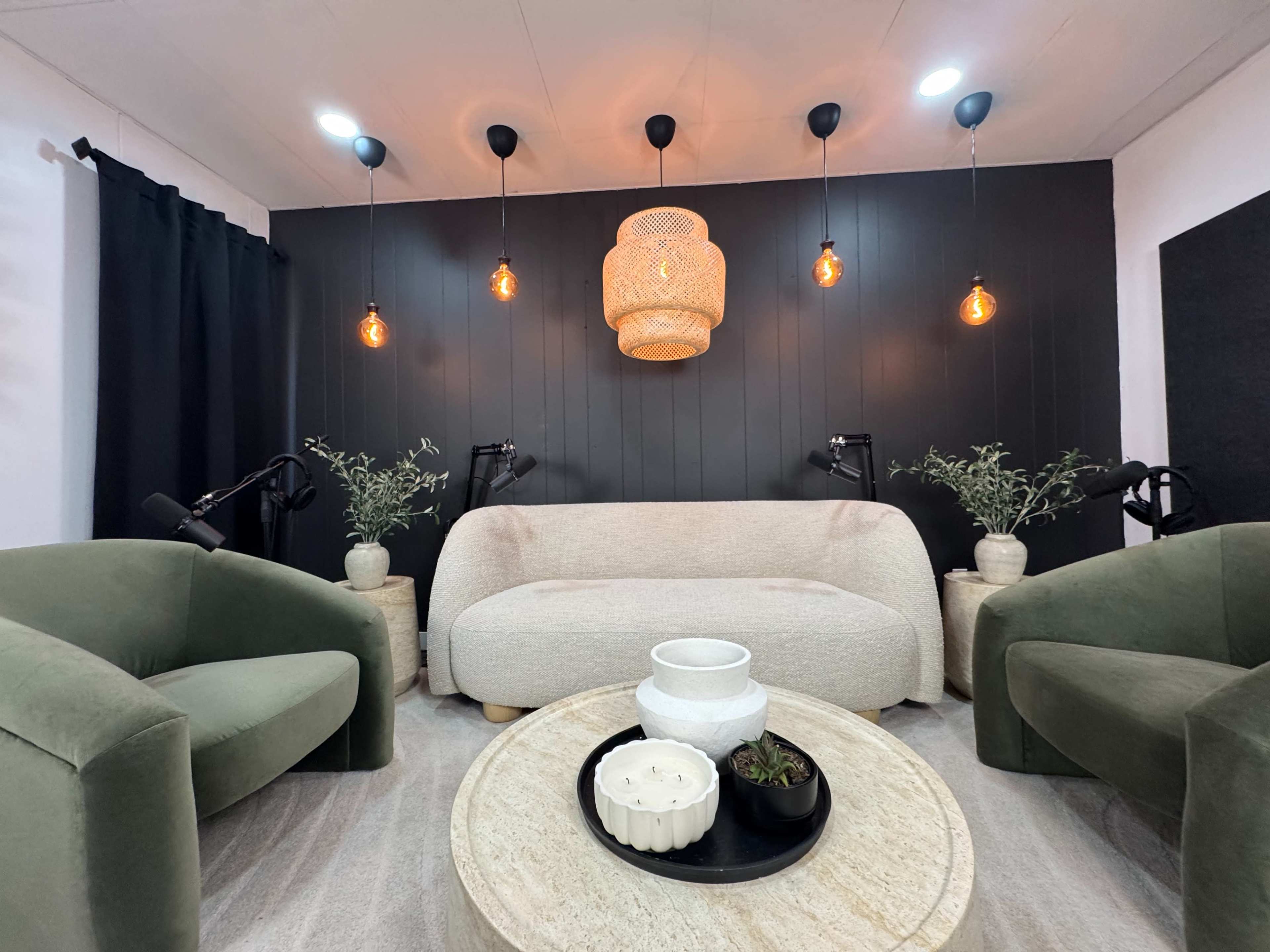 The image shows a contemporary seating area featuring a light-colored sofa, two green armchairs, a round table with decorative items, and pendant lights hanging from a black wall.