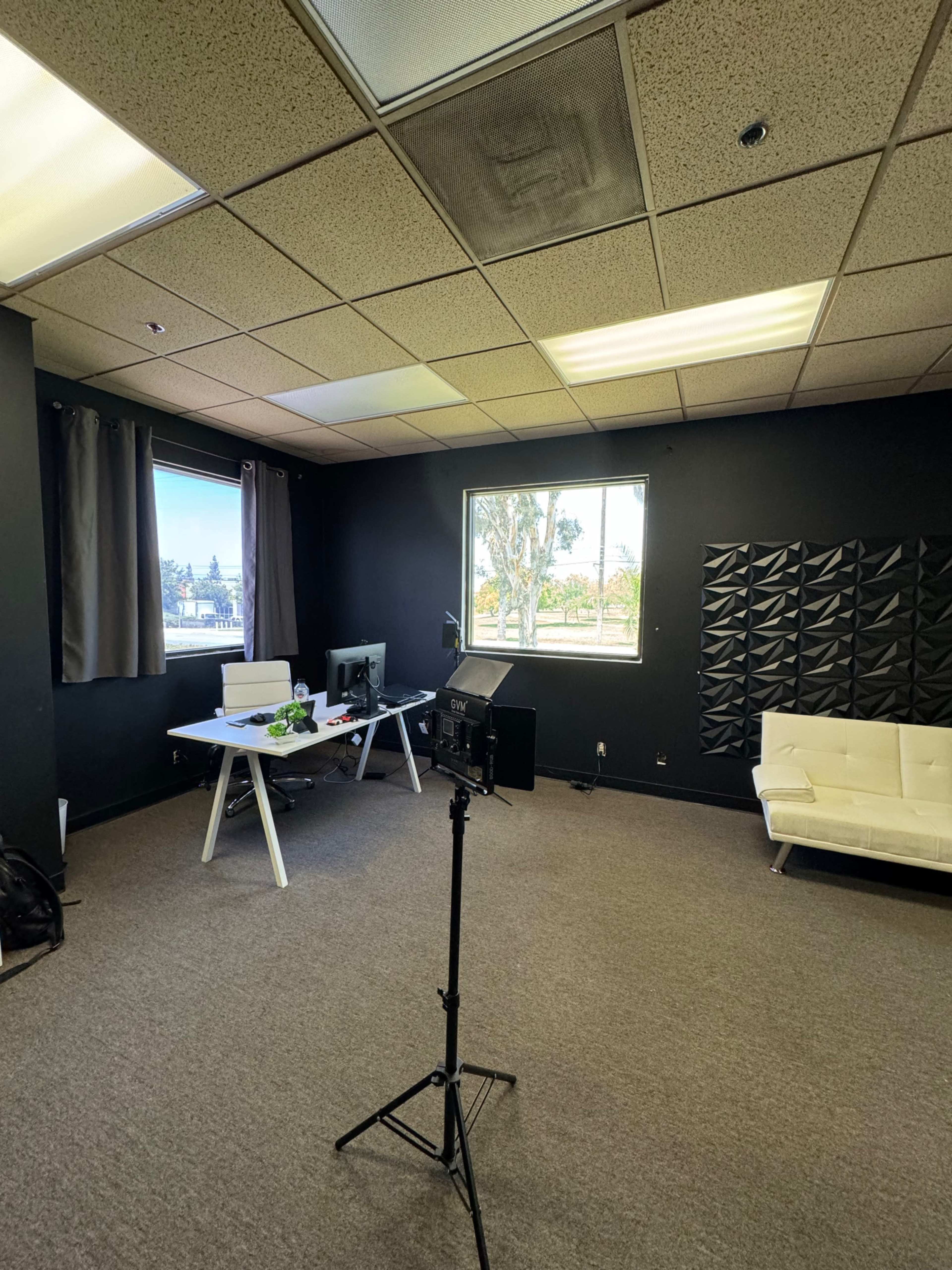 The image shows a sparsely furnished office with a black accent wall, a white desk and chair, a light-colored sofa, and a standing light on a tripod.