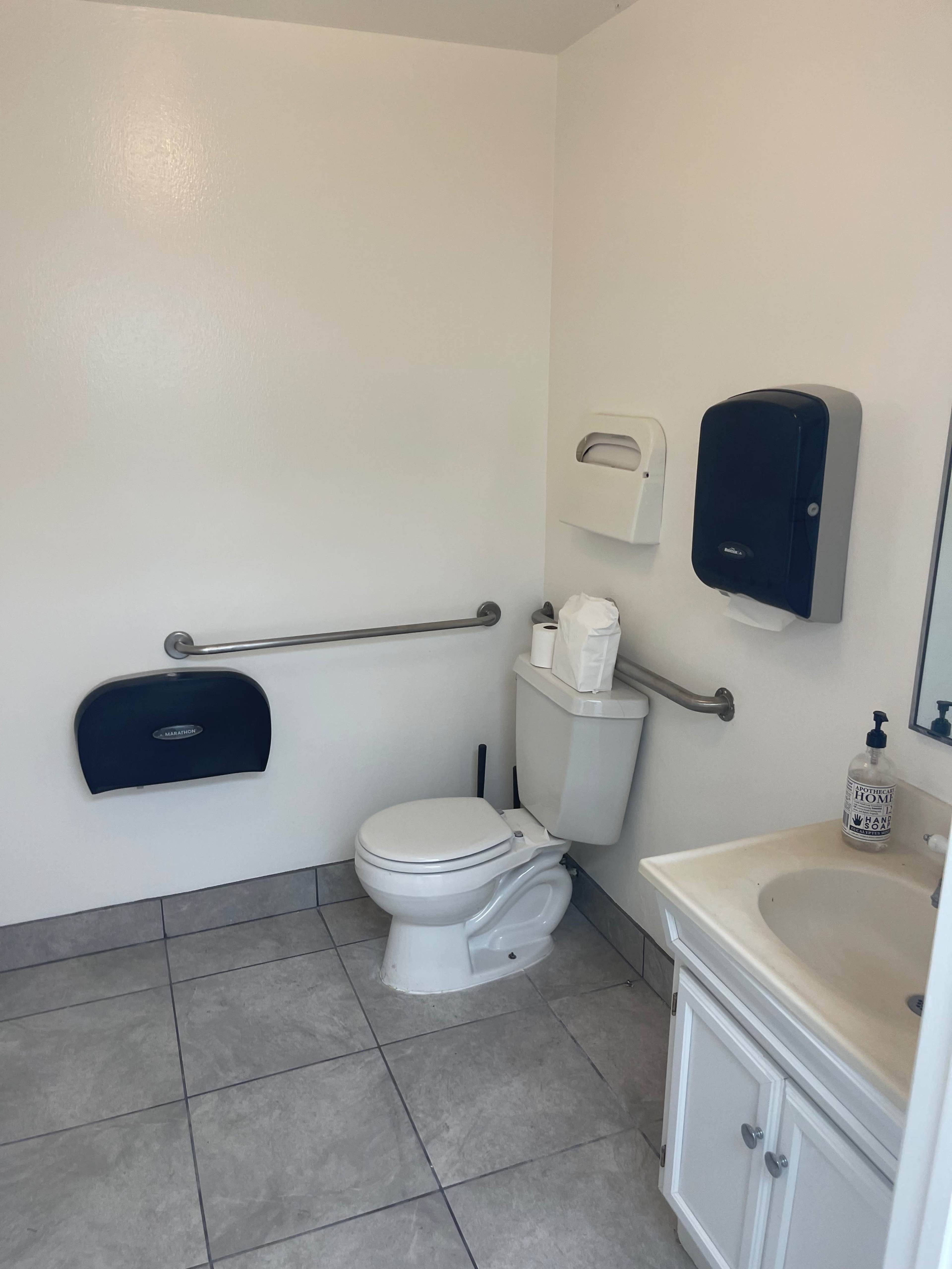 A small bathroom with a toilet, sink, and various dispensers on the wall.