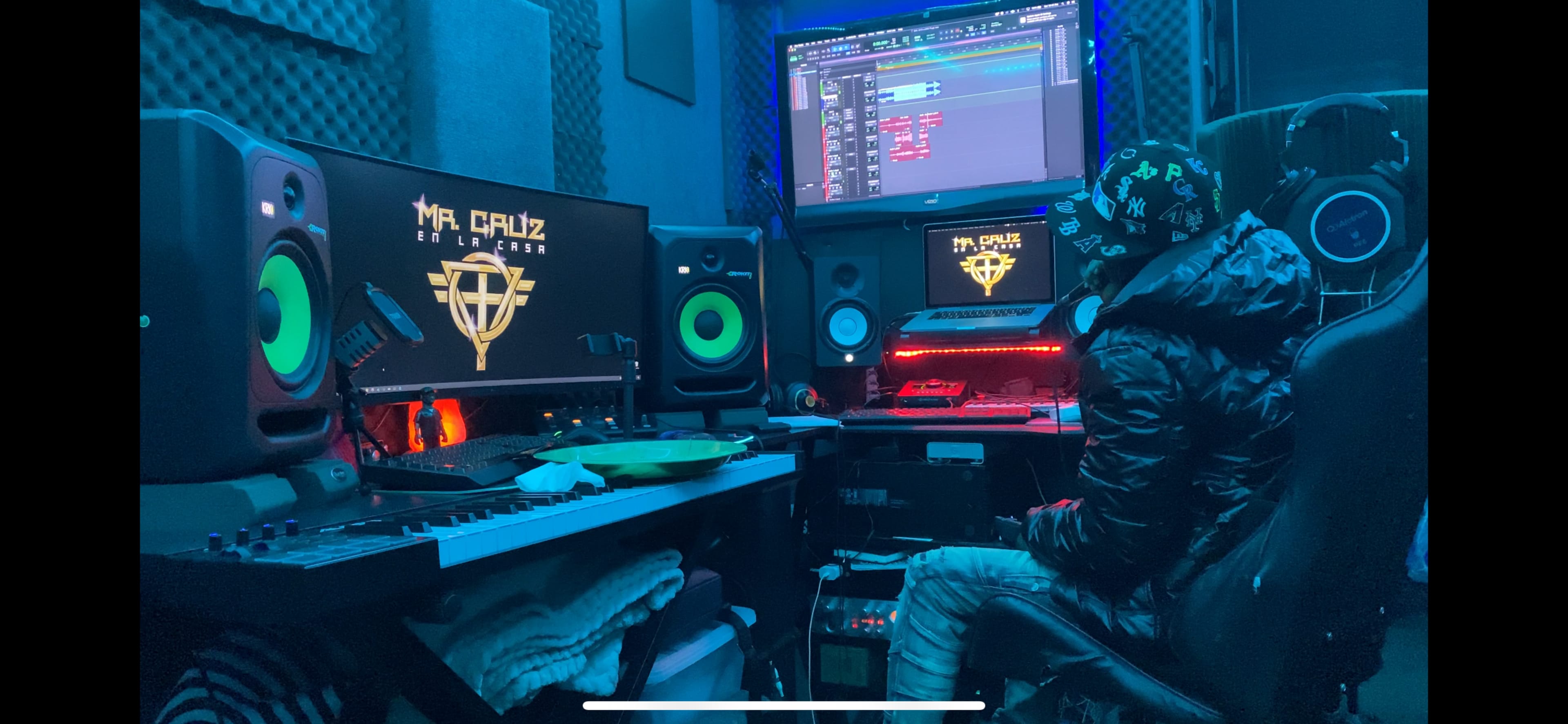 A person sits at a computer in a music production studio, surrounded by monitors, speakers, and various sound equipment.