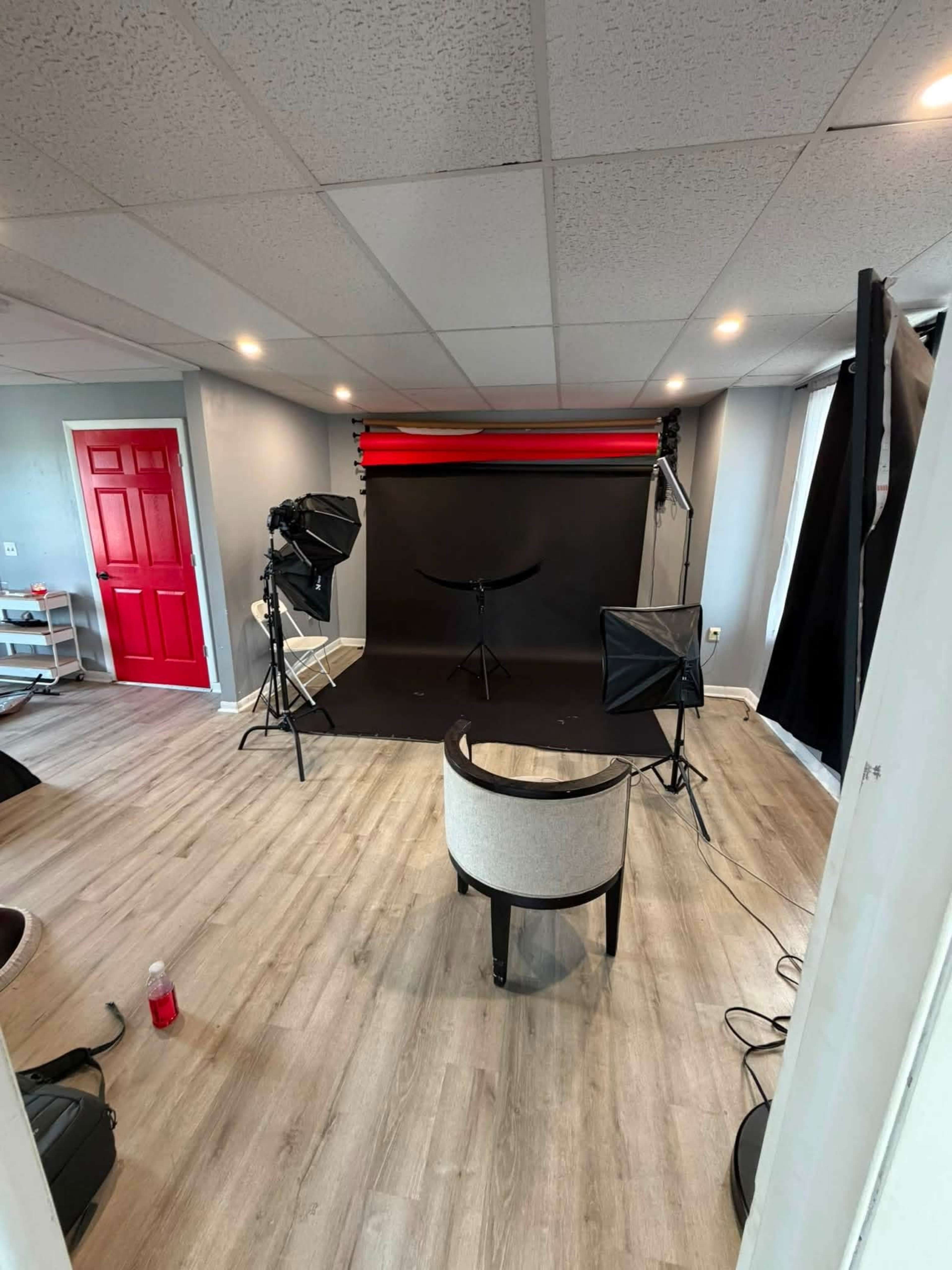 The image shows a photography studio with a red door, a black backdrop set up with lighting equipment, and a chair in the center of the room.