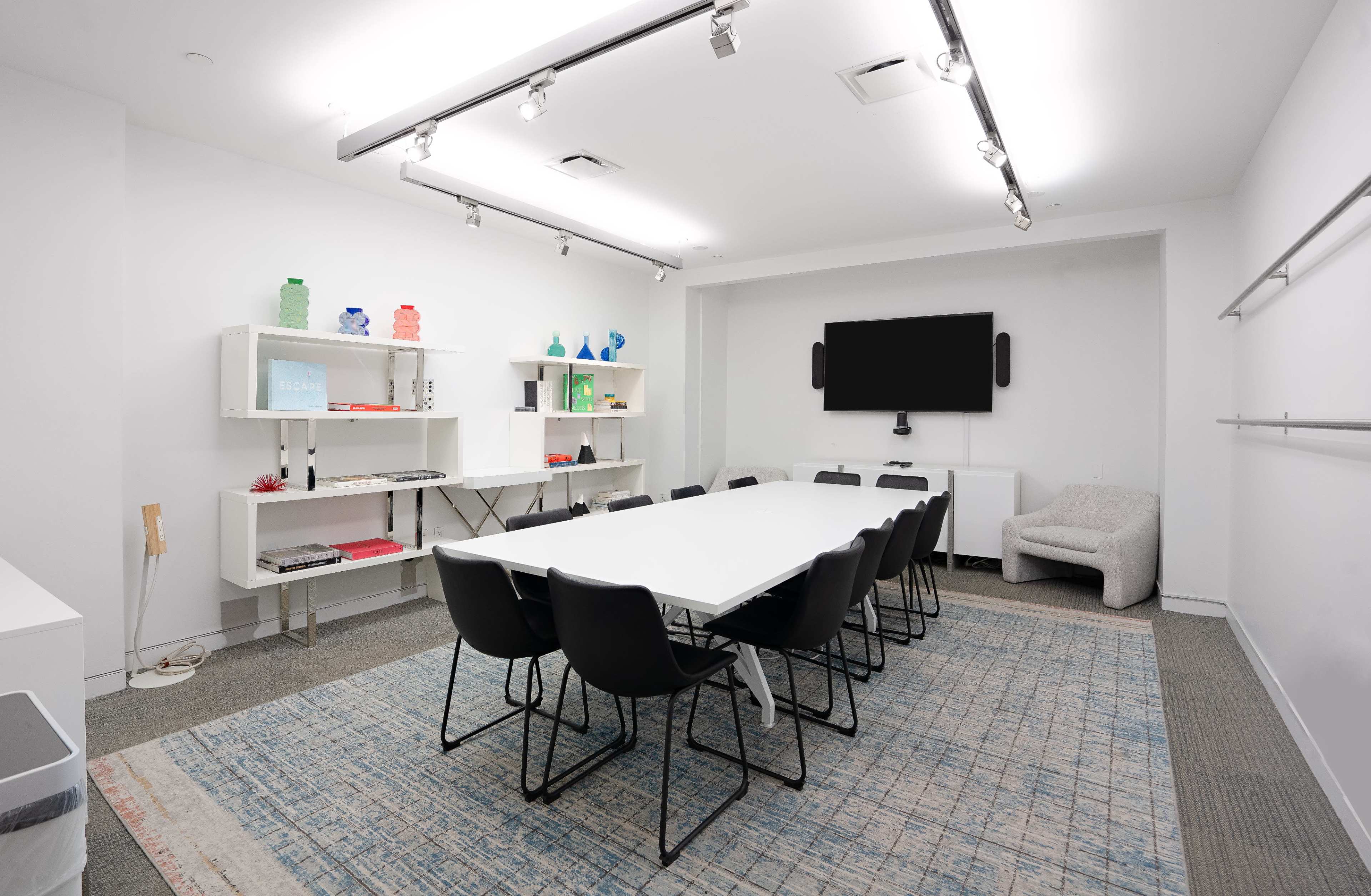 A modern conference room features a long white table surrounded by black chairs, with shelves displaying colorful decor and a wall-mounted television.