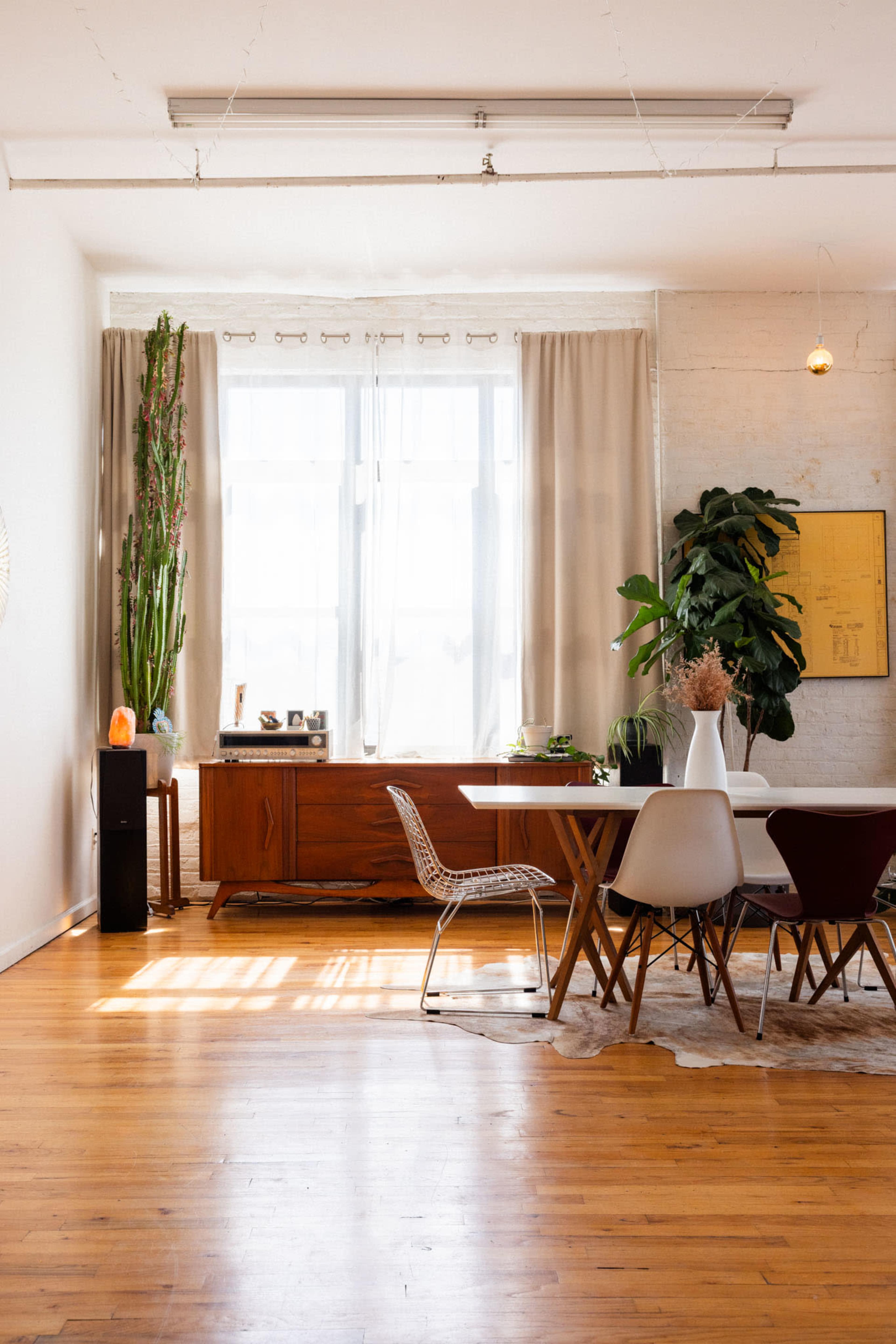 A well-lit dining area features a wooden table surrounded by chairs, a sideboard, and large windows with curtains, complemented by plants and a decorative element.