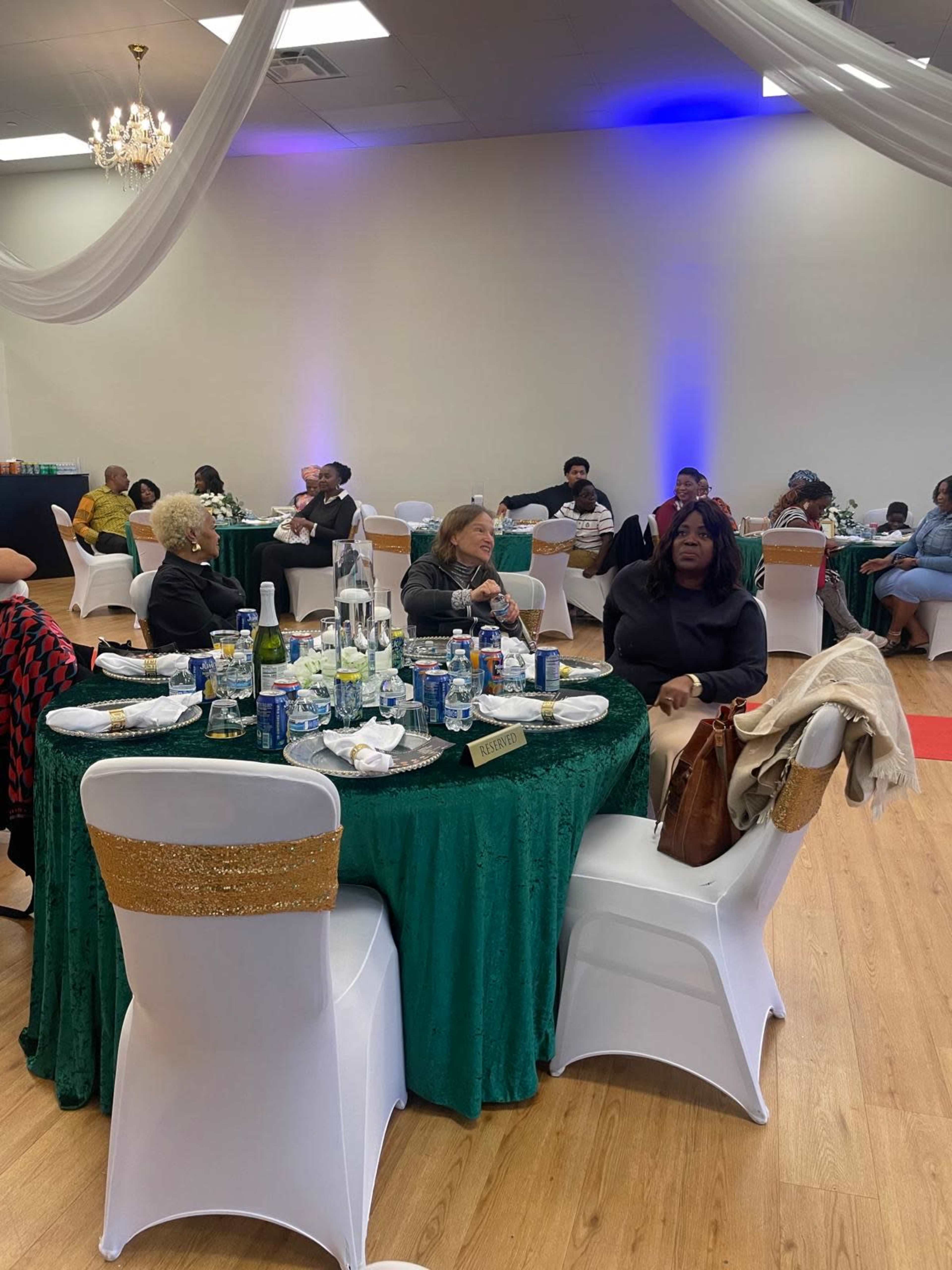 The image shows a banquet hall filled with guests seated at elegantly decorated tables covered in green tablecloths, with plates and drinks scattered on the tables.