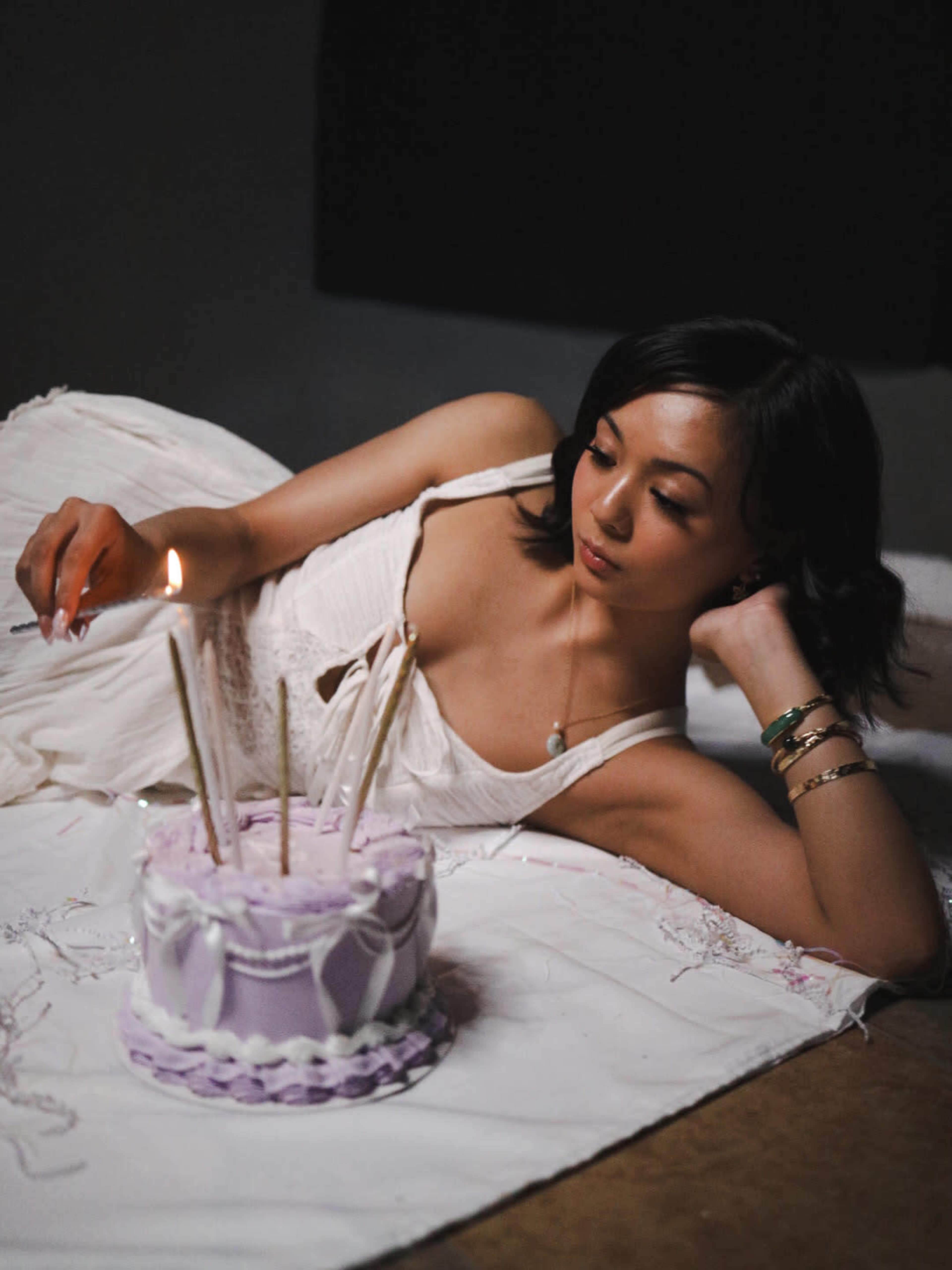 A person is lying beside a decorated birthday cake with lit candles, wearing a white dress and looking thoughtfully at the cake.