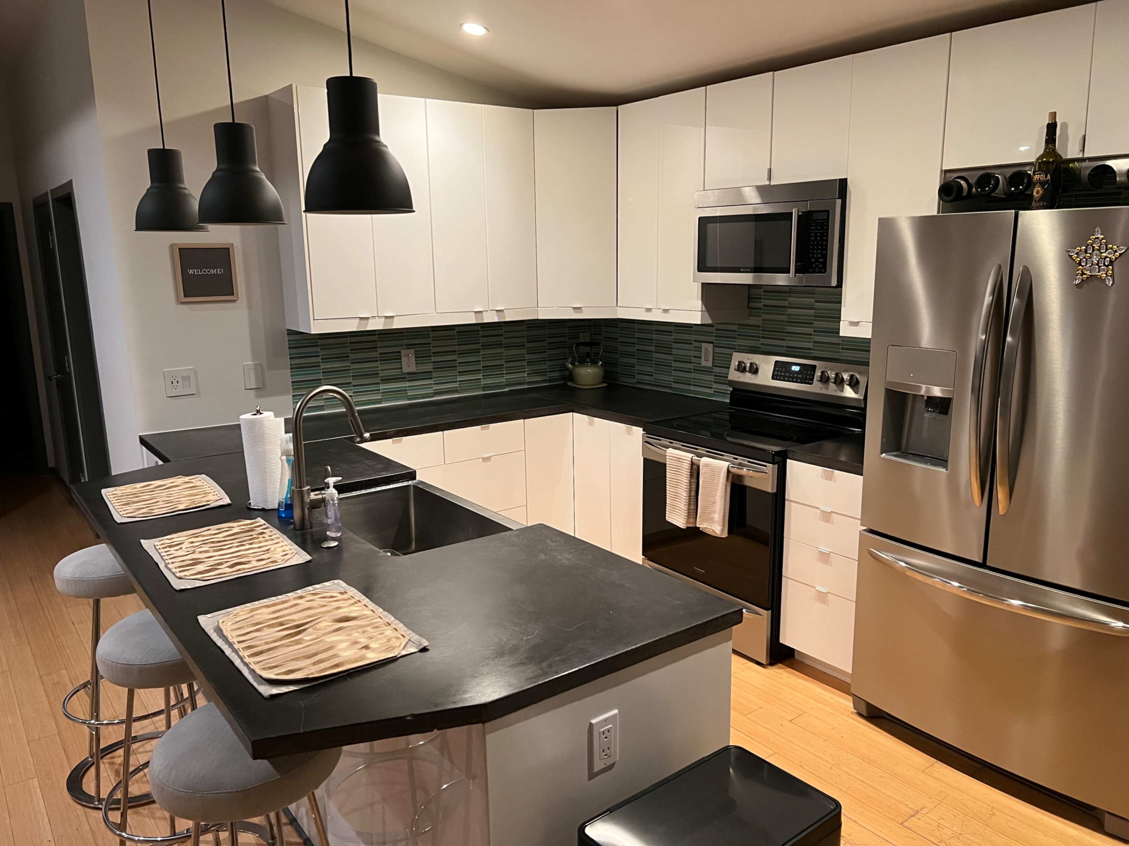 A modern kitchen with white cabinets, stainless steel appliances, and a black countertop, complemented by bar stools and pendant lighting.