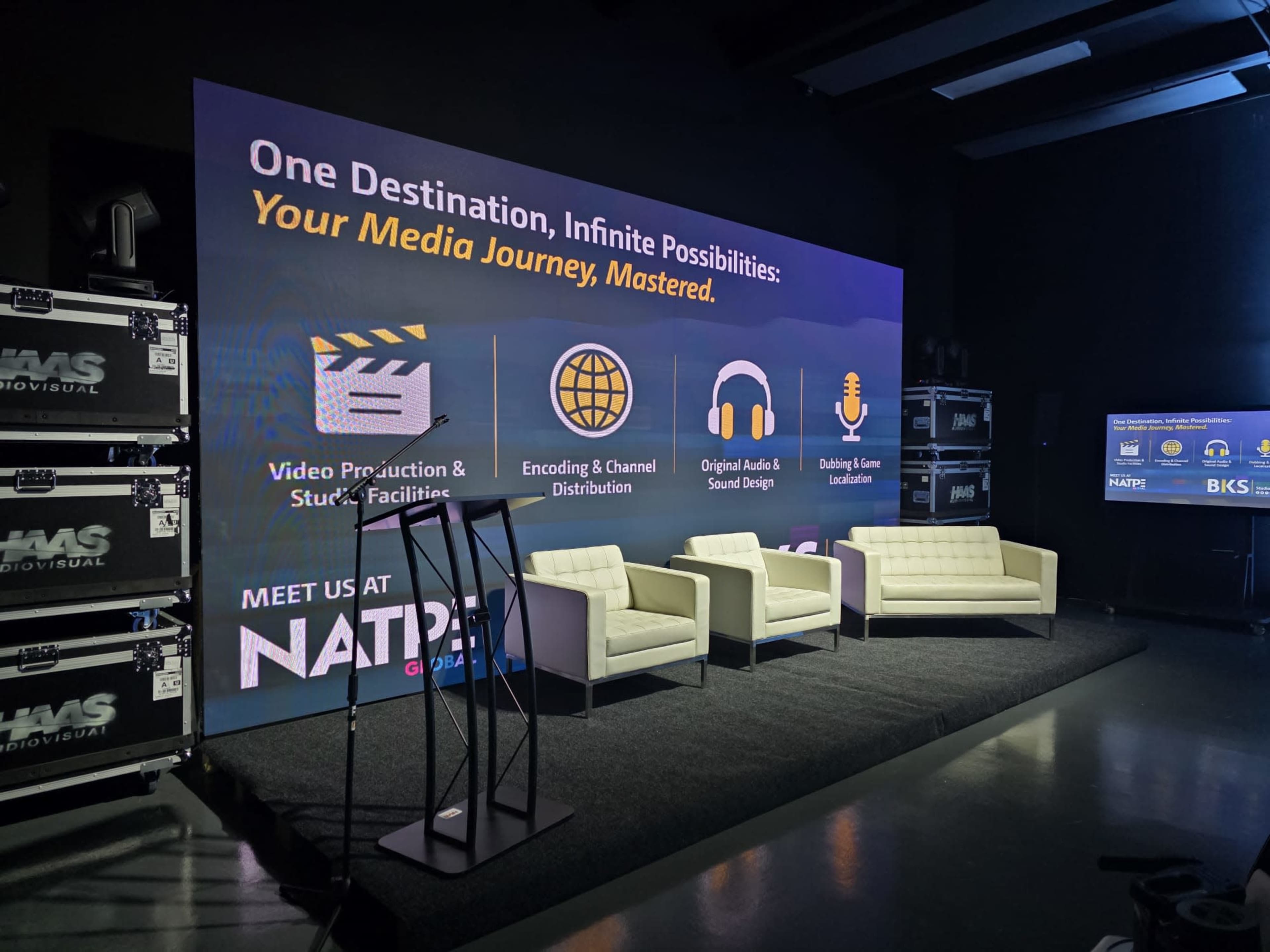 The image shows a stage setup featuring a large backdrop with marketing text, two white couches, and a microphone stand in front.