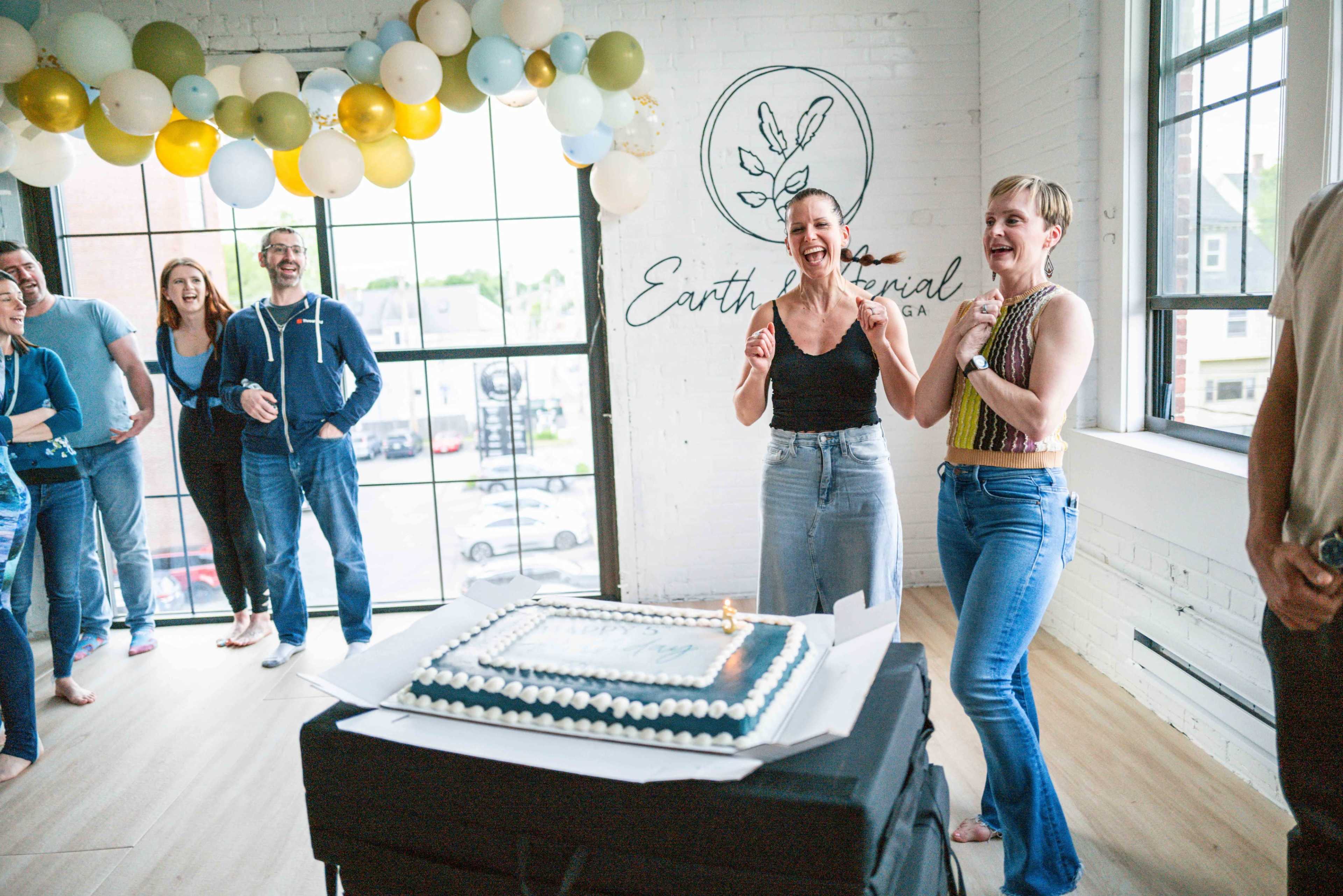 A group of people celebrate with laughter and smiles around a cake in a brightly lit room adorned with balloons.