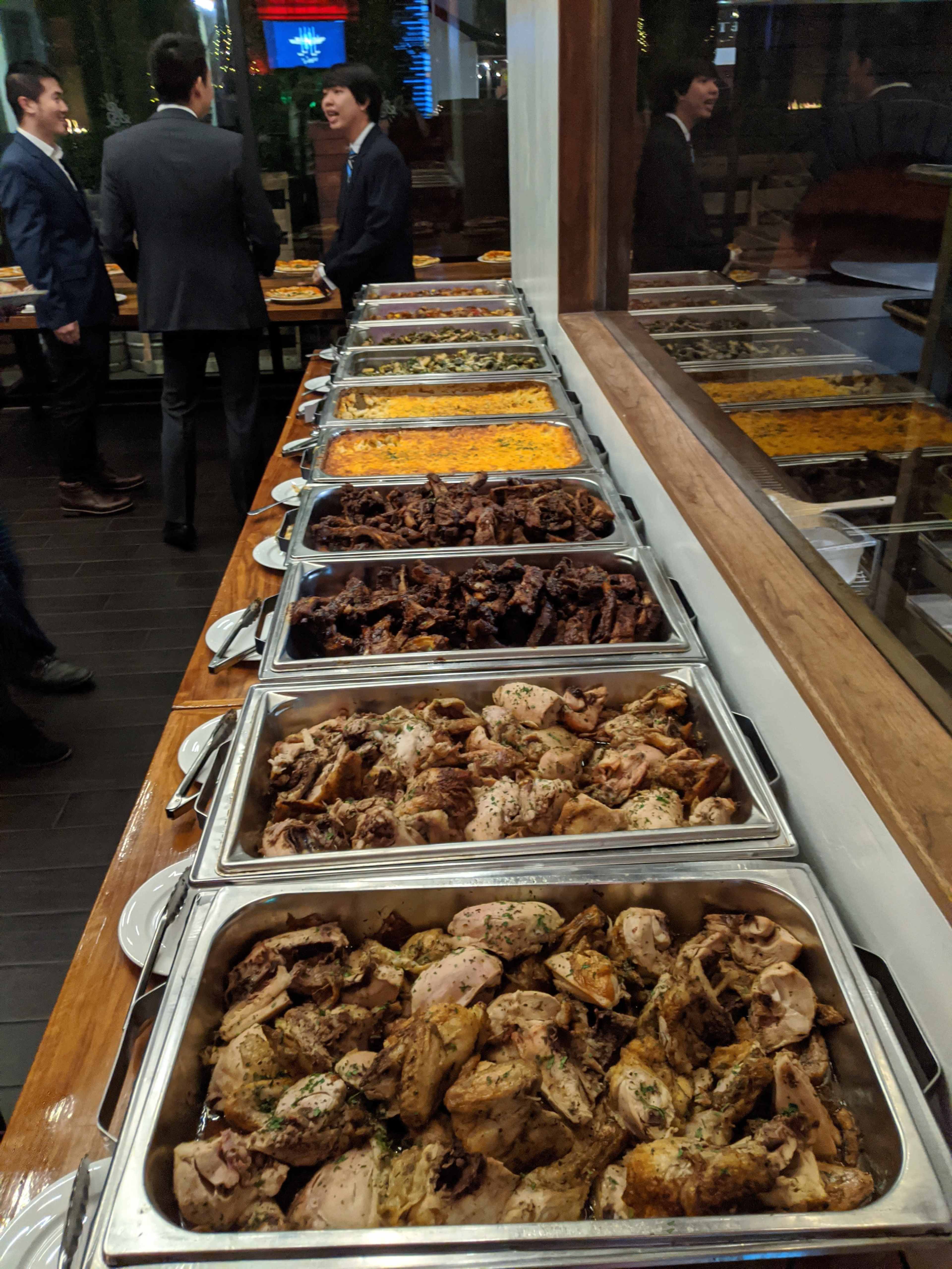 A long table is set with multiple trays of various cooked meats, including chicken and beef, alongside sides like rice and other dishes, while people converse in the background.