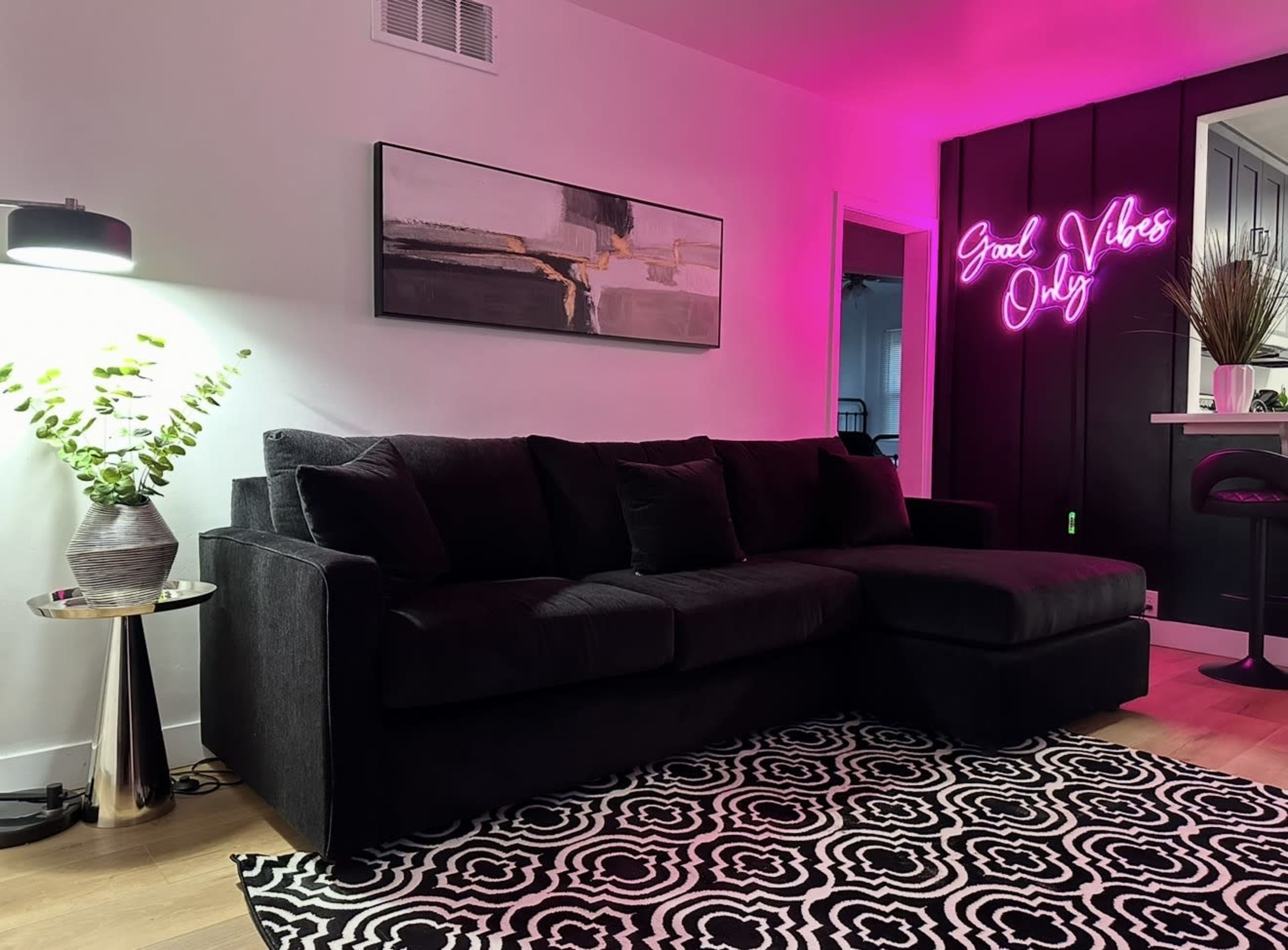 A black sectional sofa with decorative pillows is situated next to a metallic side table, illuminated by a lamp, while a neon sign reading "Good Vibes Only" casts a pink glow against the wall.