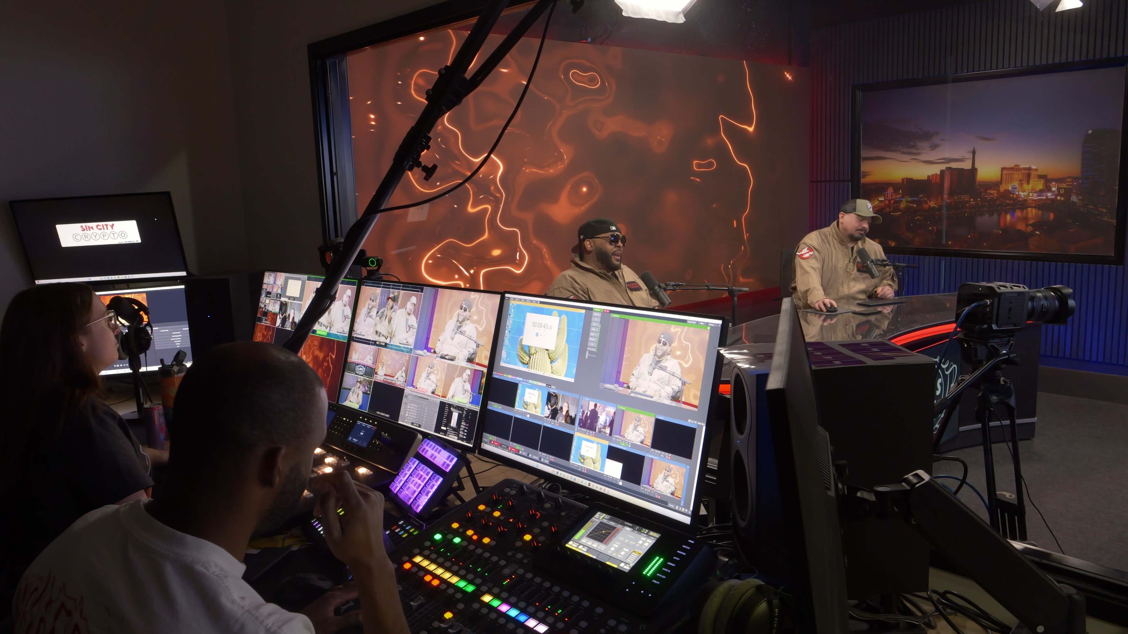 The image shows a busy television studio control room with multiple screens and a crew operating equipment while two hosts are visible on a set in the background.