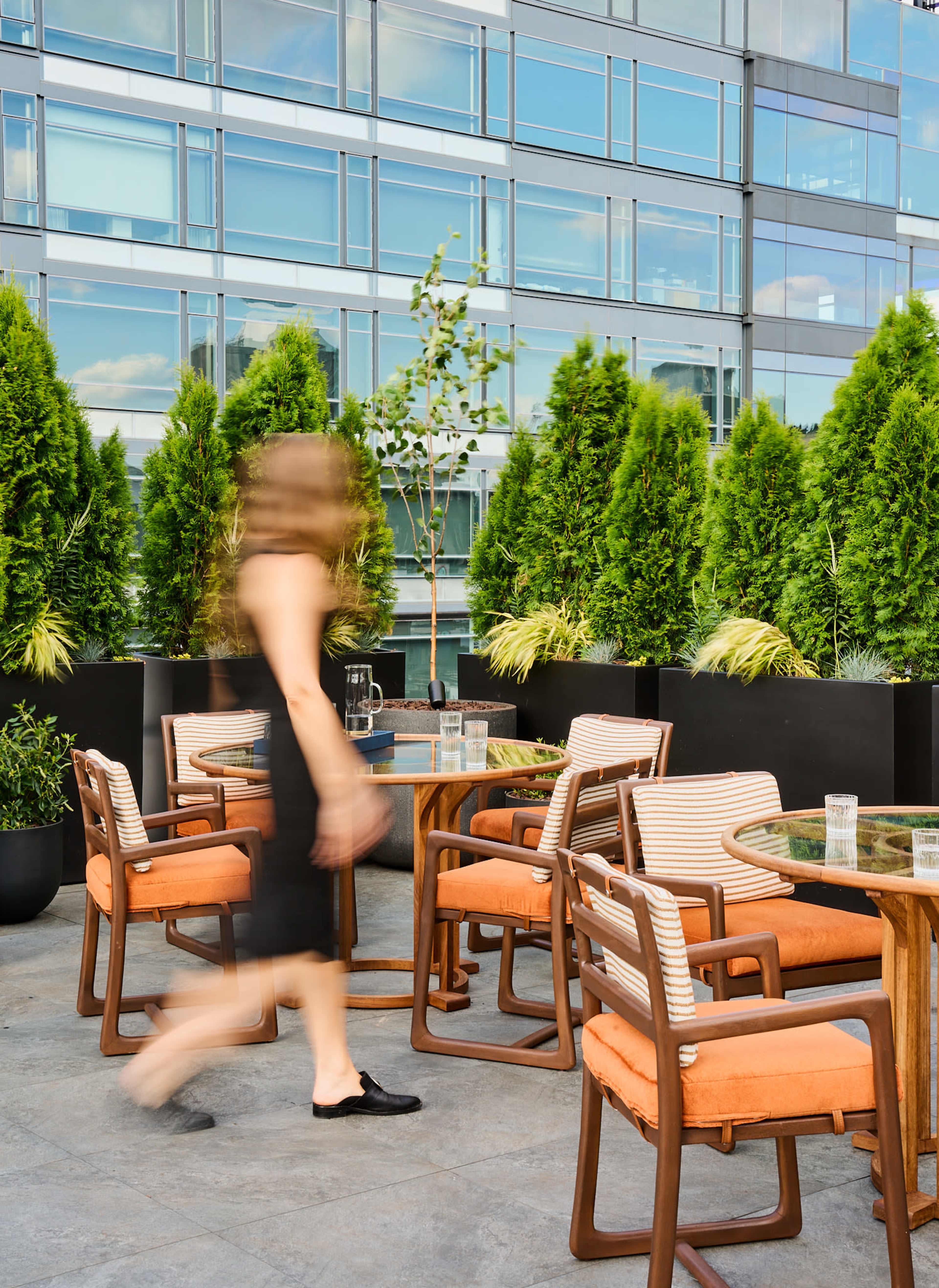 A woman walks past empty tables and chairs on a modern outdoor terrace surrounded by greenery and glass walls.