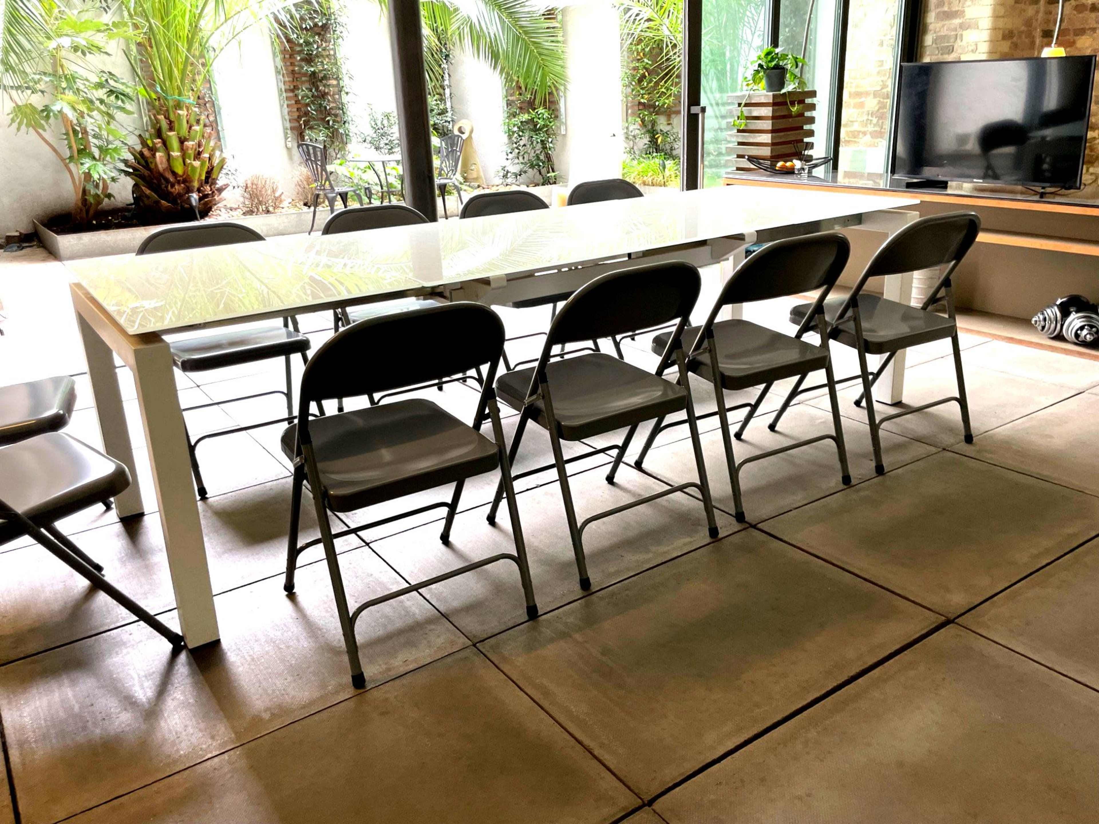 A long, white dining table with several gray folding chairs is positioned in a room with large windows and potted plants.