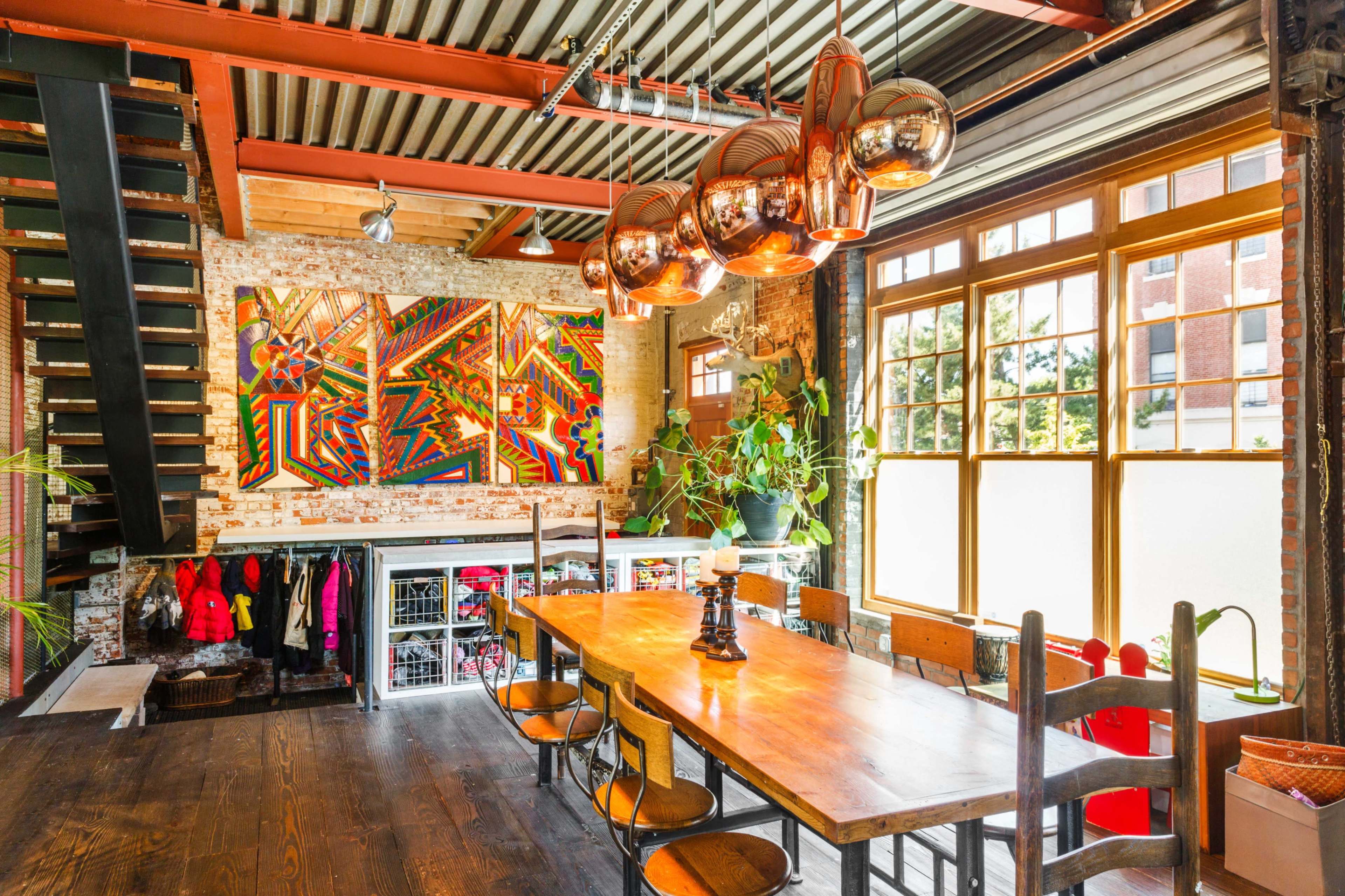 The image shows a spacious industrial-style dining area with a large wooden table, copper pendant lights, and a colorful artwork on the brick wall, surrounded by large windows.