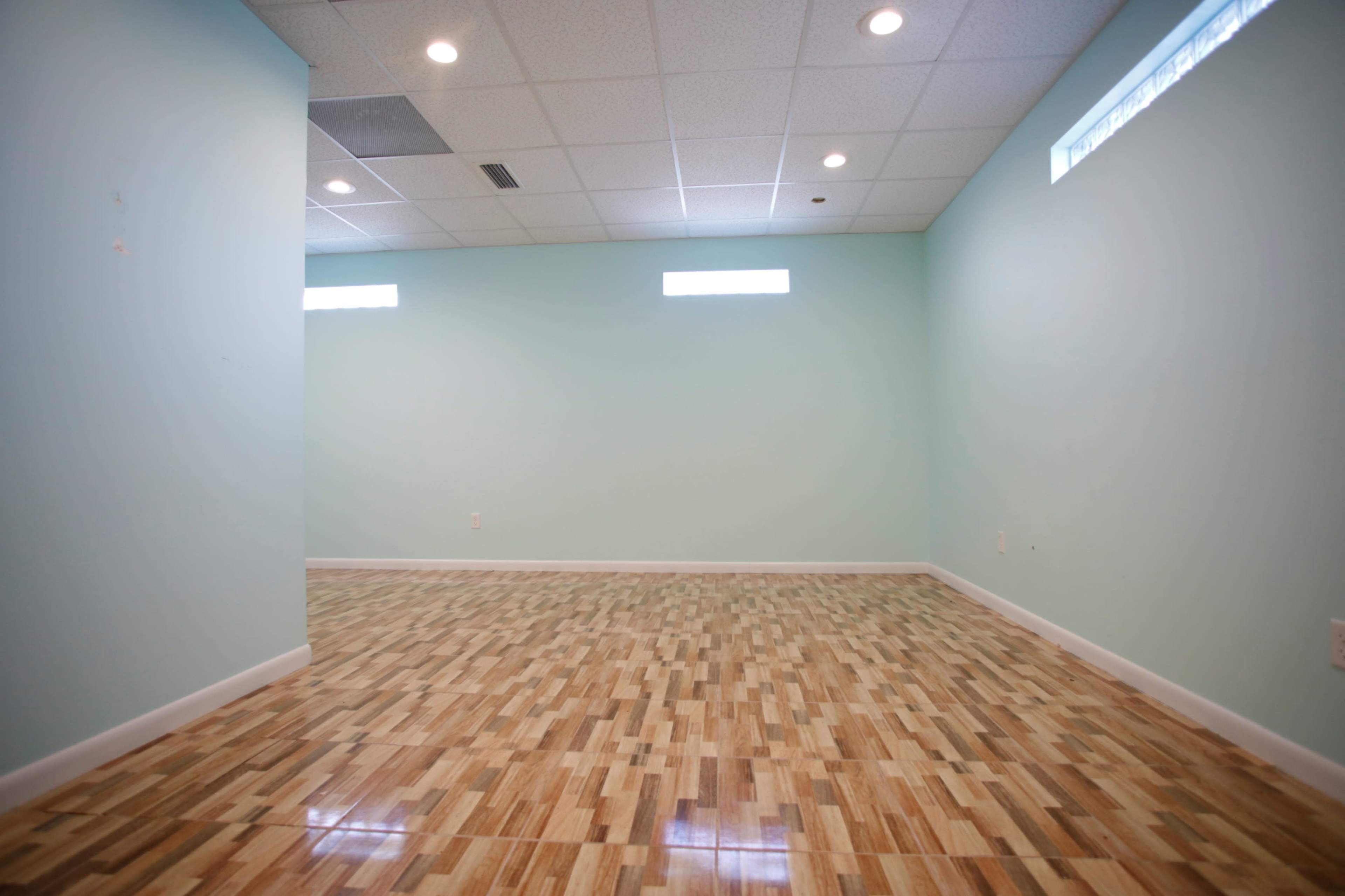 The image shows a vacant room with light green walls and a polished wooden floor, illuminated by overhead lights and small windows.