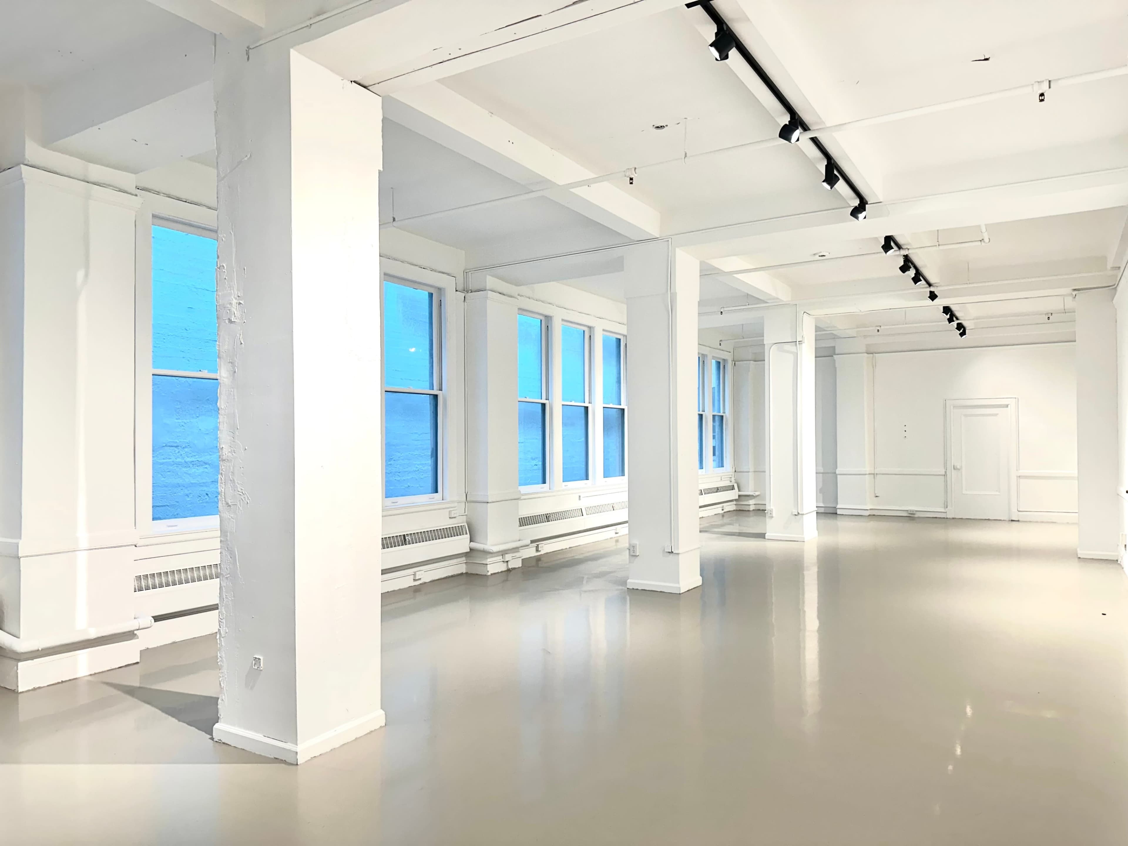 The image shows a spacious, bright room with white walls and large windows providing a view of a body of water.