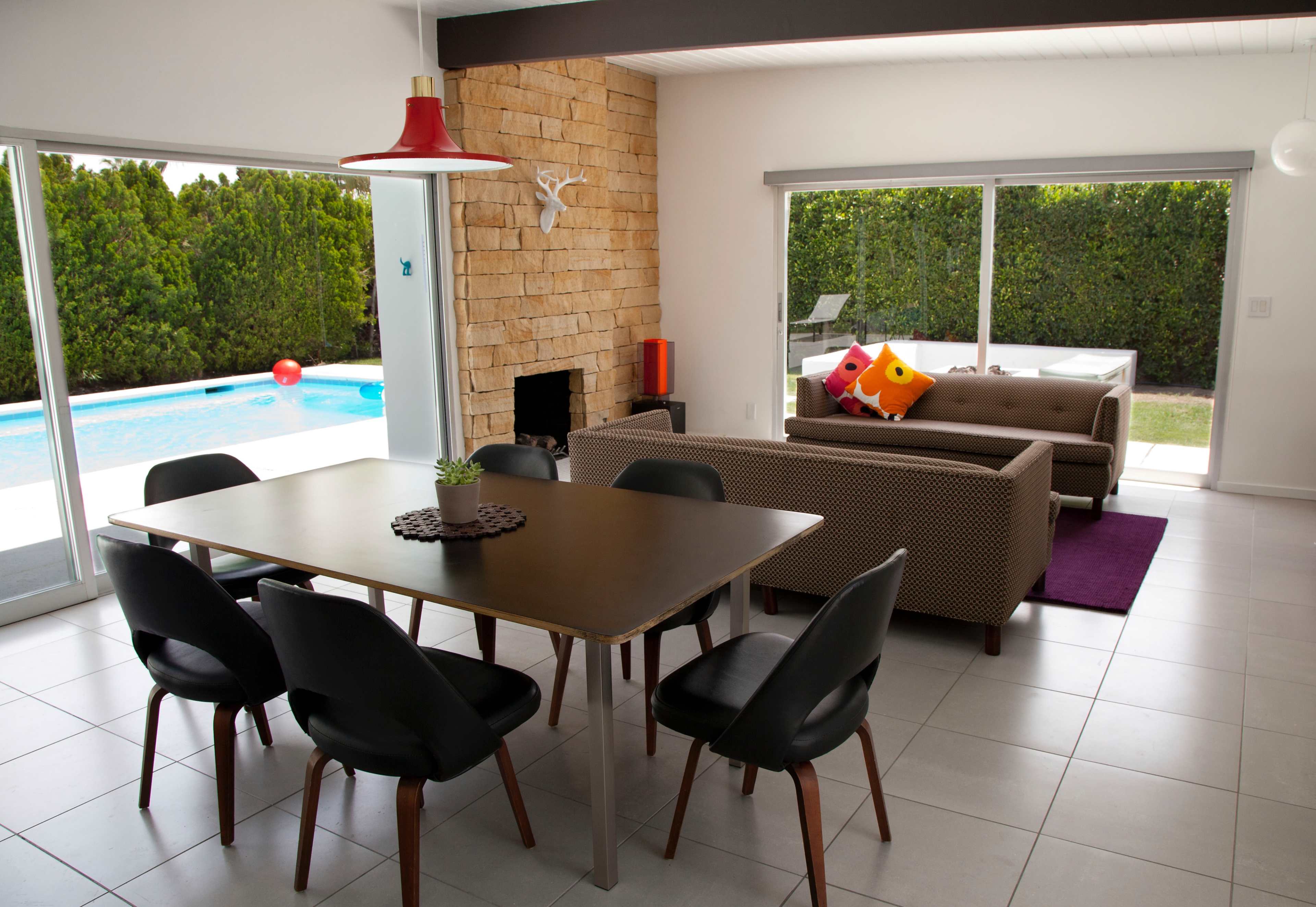 The image shows a modern living room and dining area with a view of a pool and greenery outside.