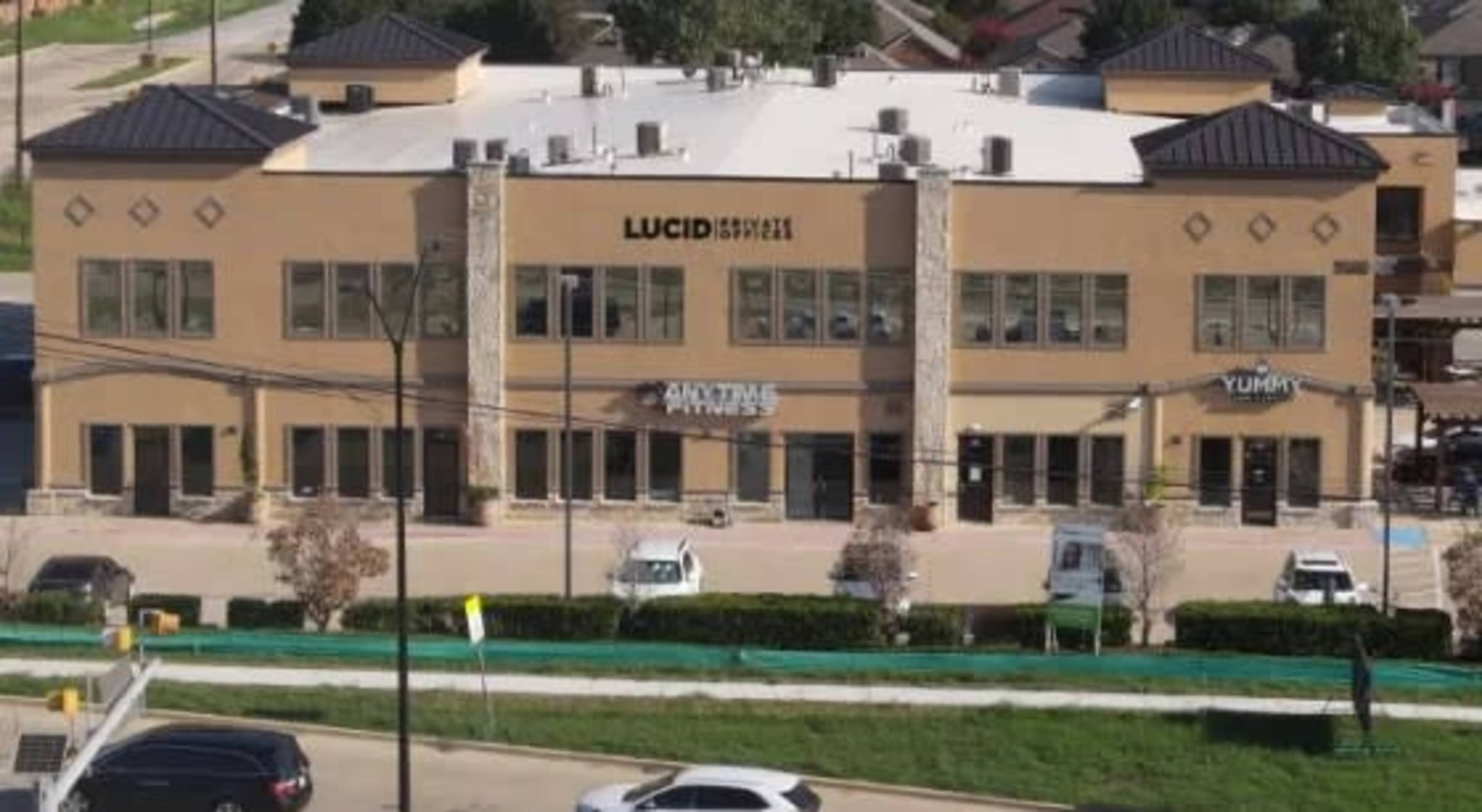 The image shows a two-story commercial building featuring businesses such as Lucid, Anytime Fitness, and Yummy, with parking spaces in front.