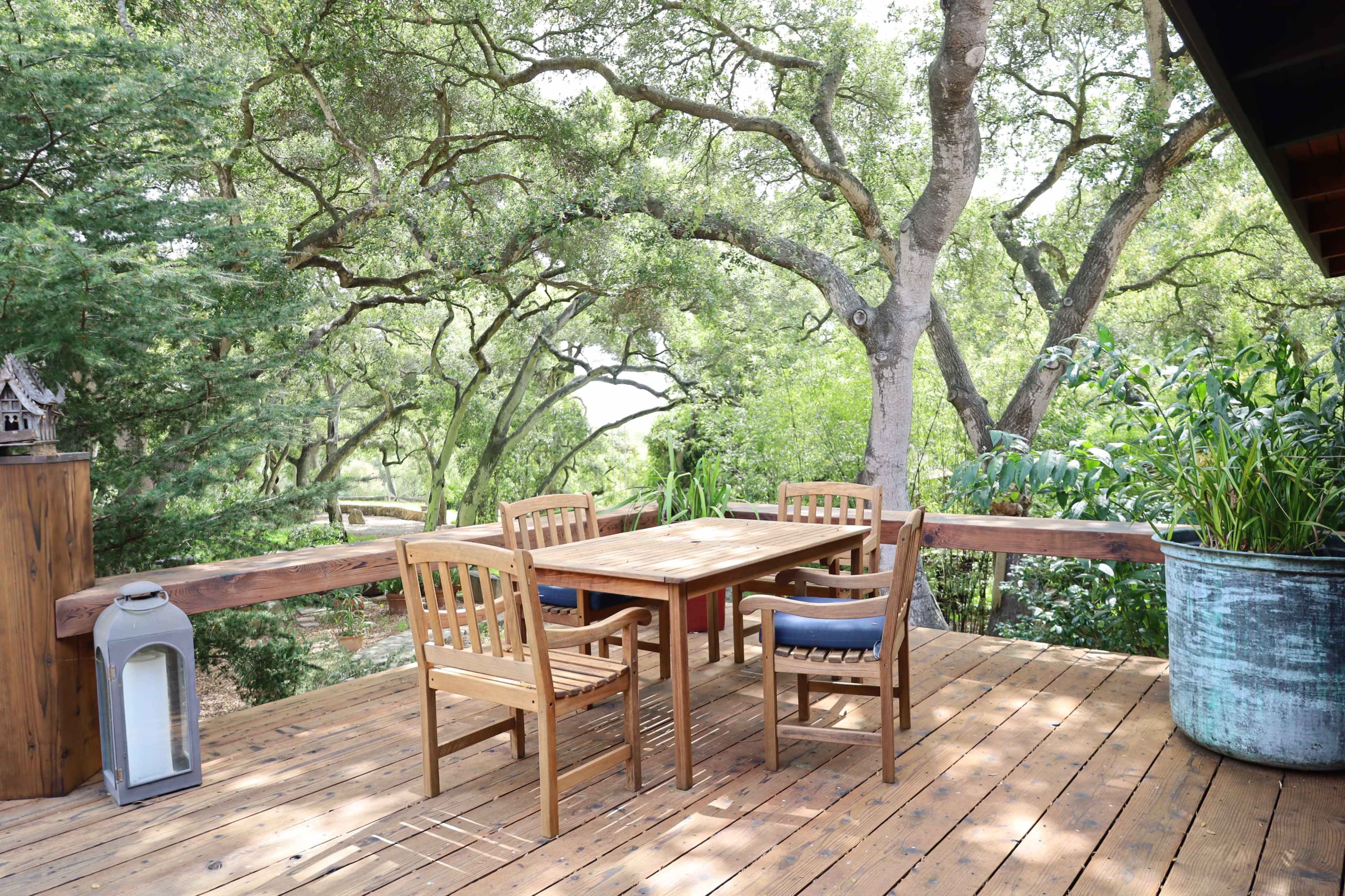 A wooden deck features a table and four chairs surrounded by trees and greenery.