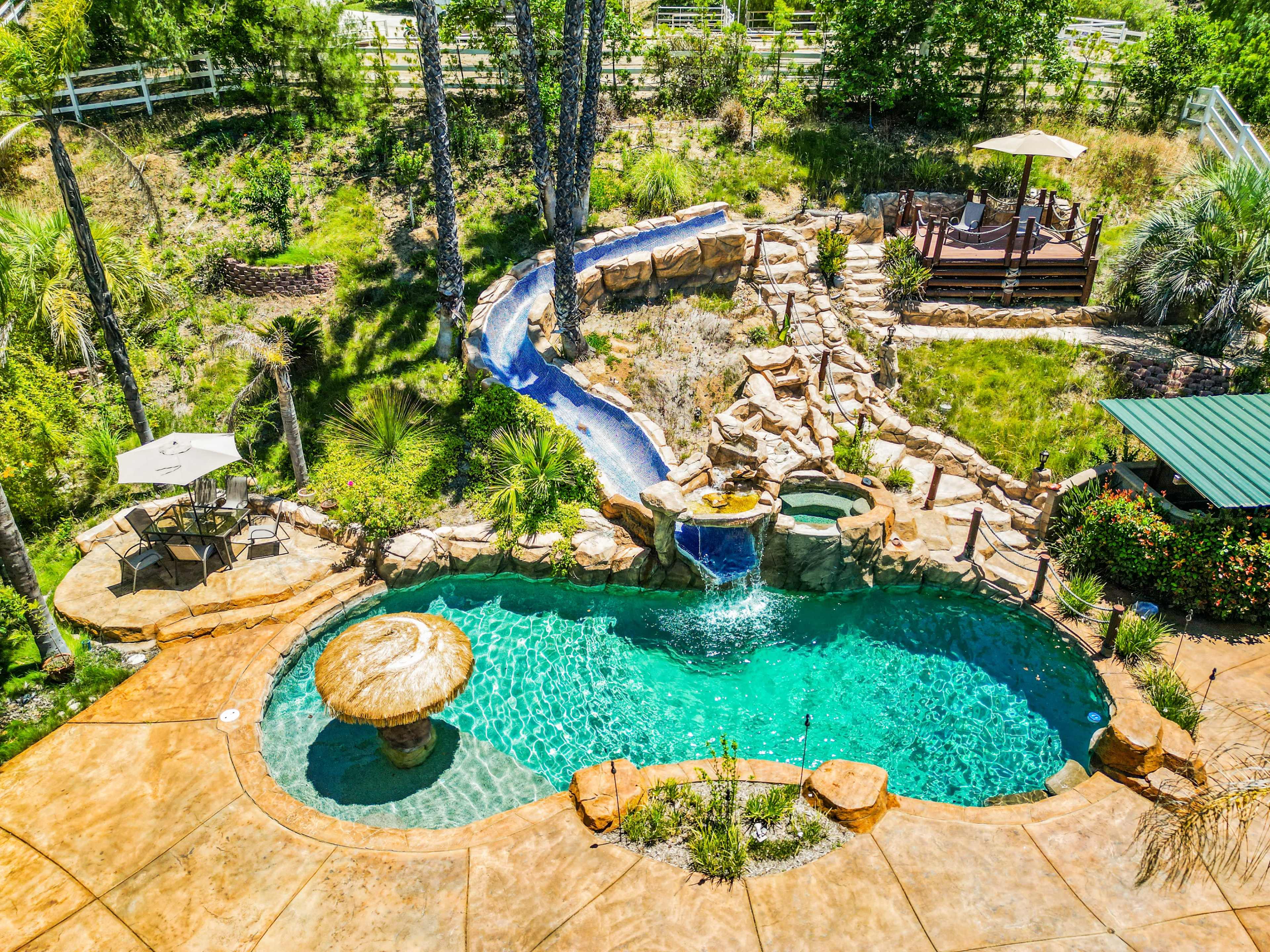 The image depicts a landscaped backyard featuring a swimming pool with a rock waterfall, a stone pathway leading to a shaded seating area, and a waterslide winding down into the pool.
