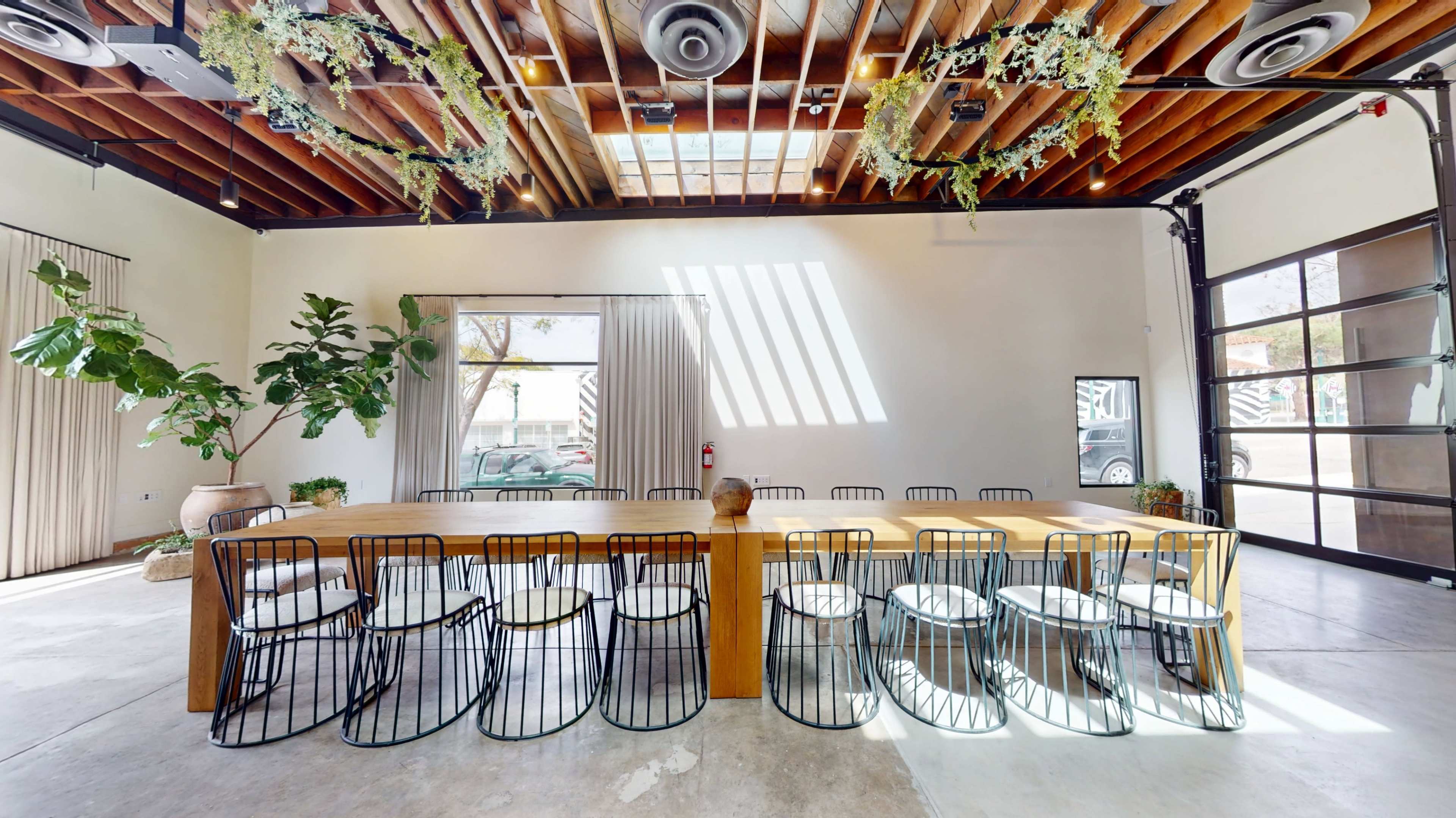 A spacious dining area features a long wooden table surrounded by black wire chairs, with large windows allowing natural light to stream in and plants decorating the space.