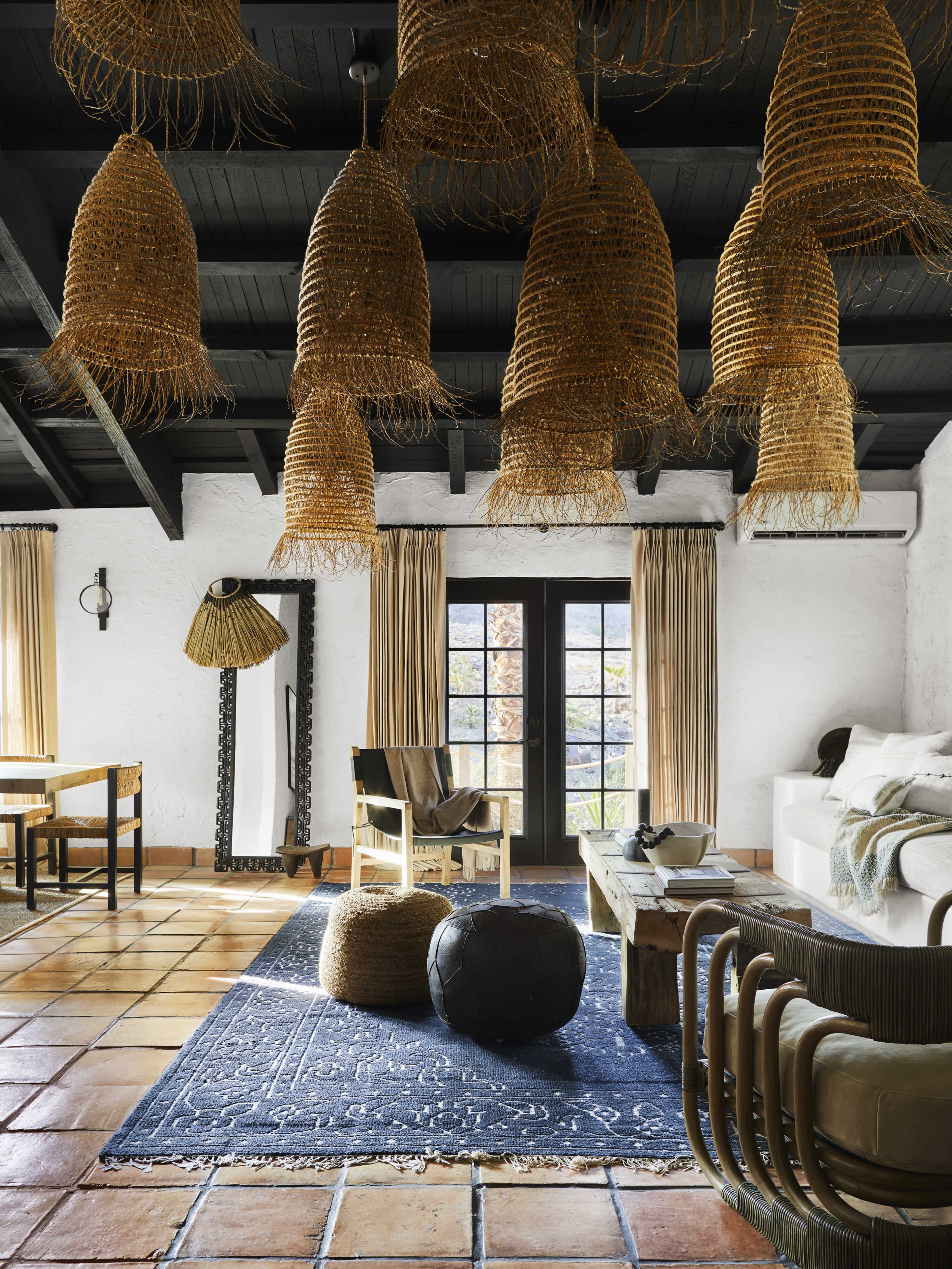 A spacious living room features textured woven light fixtures, a large patterned rug, and a mix of wooden and upholstered furniture against a backdrop of white walls and terracotta tile flooring.