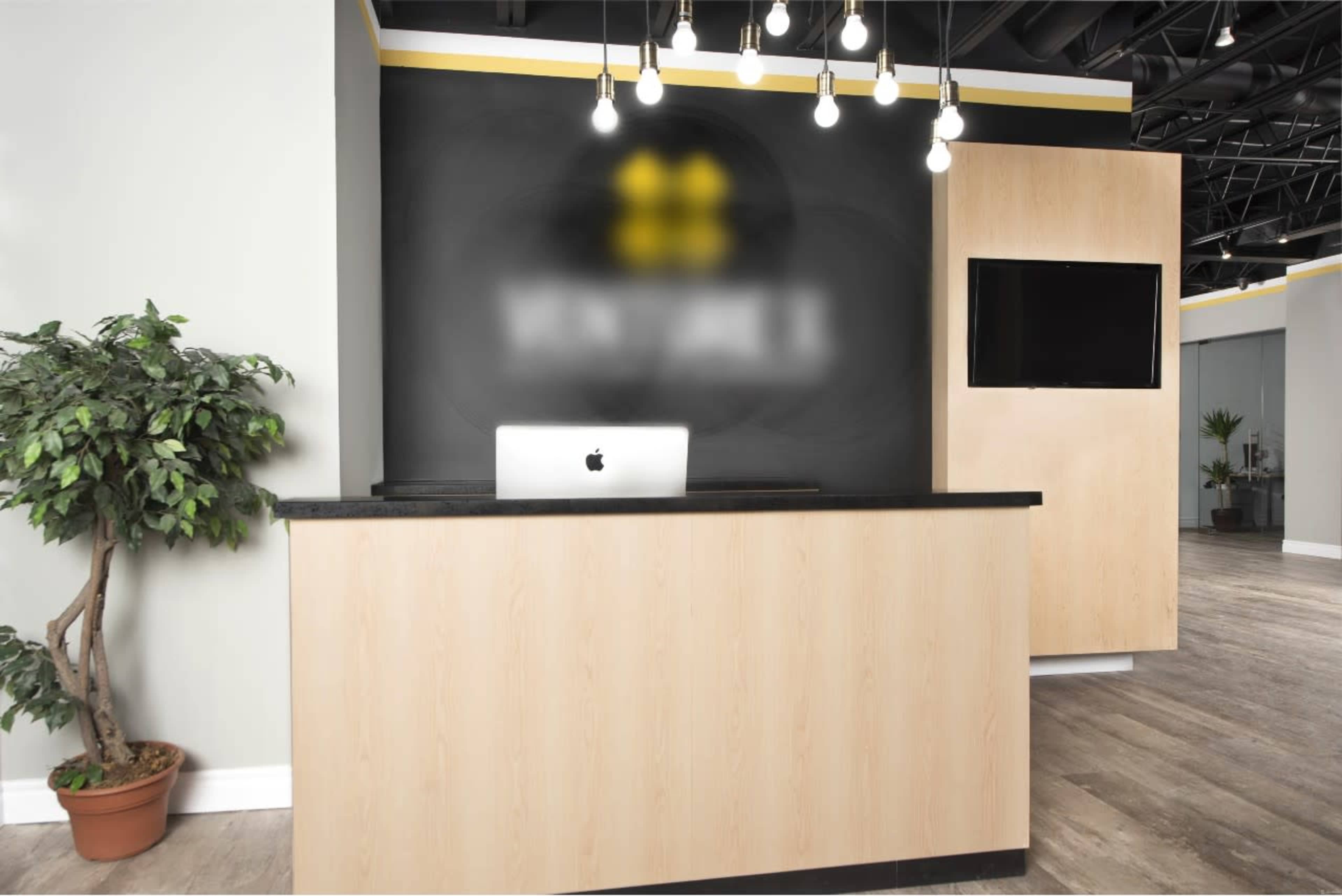 A sleek reception desk made of light wood with a black countertop, featuring a computer on it, set against a dark wall with decorative lighting and a television.