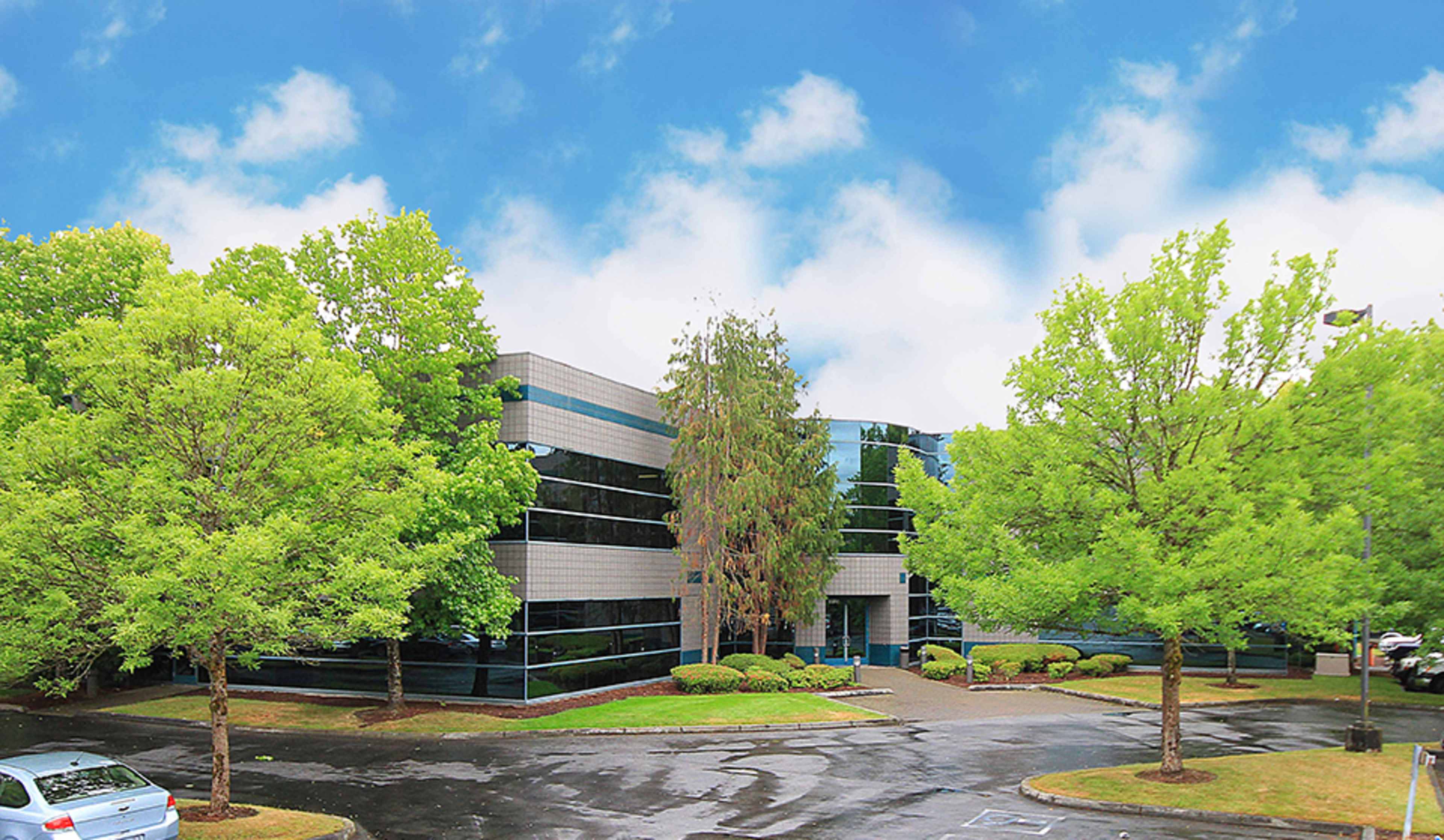A modern office building surrounded by green trees and a wet parking lot under a blue sky.